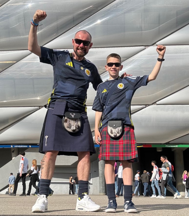 Iain and Aleks dressed in Scotland shirts and kilts, with arms raised to pose for photo