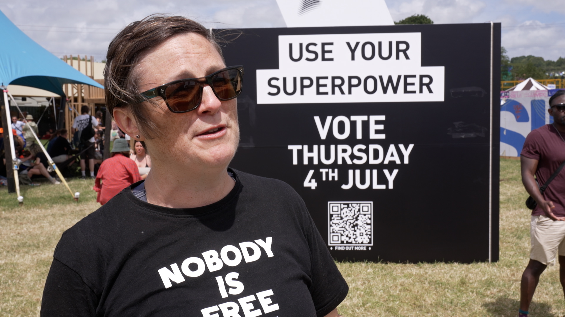 Festival-goer Anne Langford speaks in front of a black giant ballot box at Glastonbury which features the message 'Use Your Superpower. Vote Thursday 4th July'