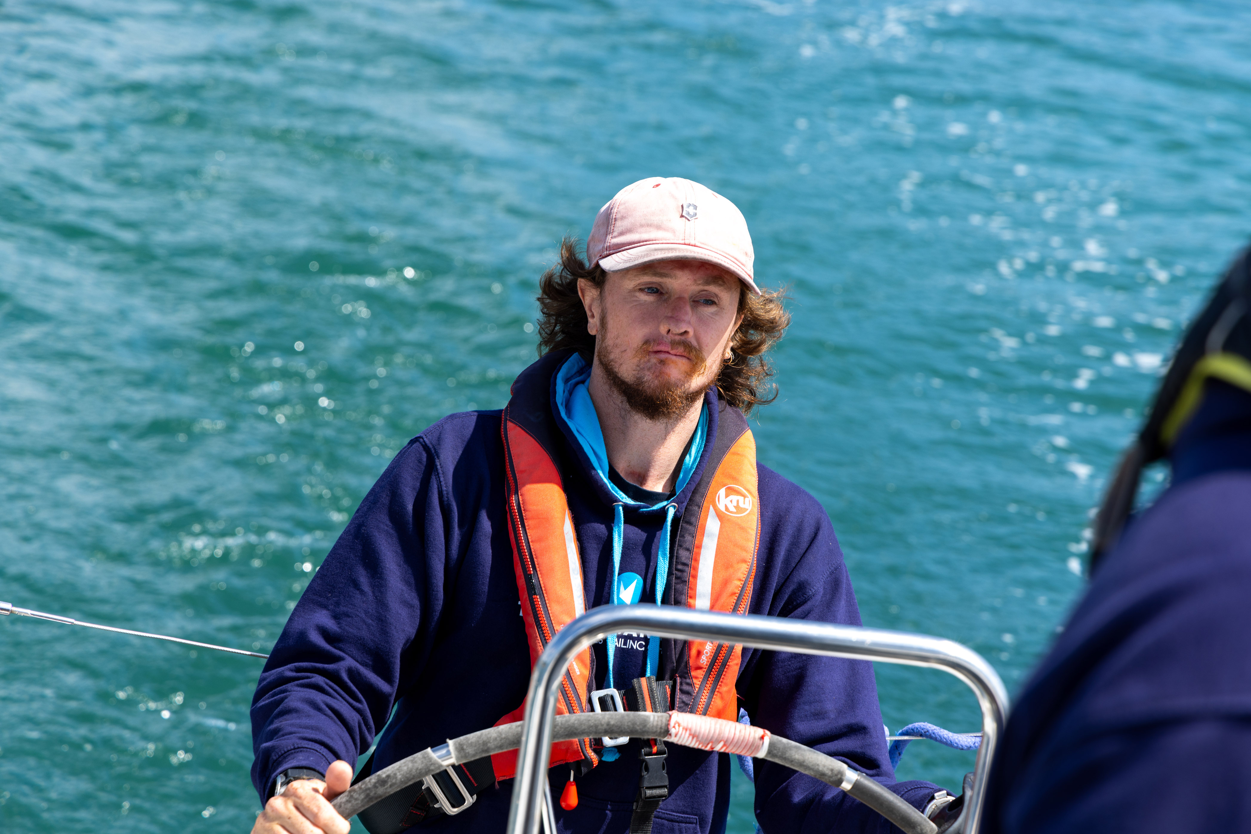 Craig Wood standing at the helm of a sailing boat wearing a life jacket and a hat