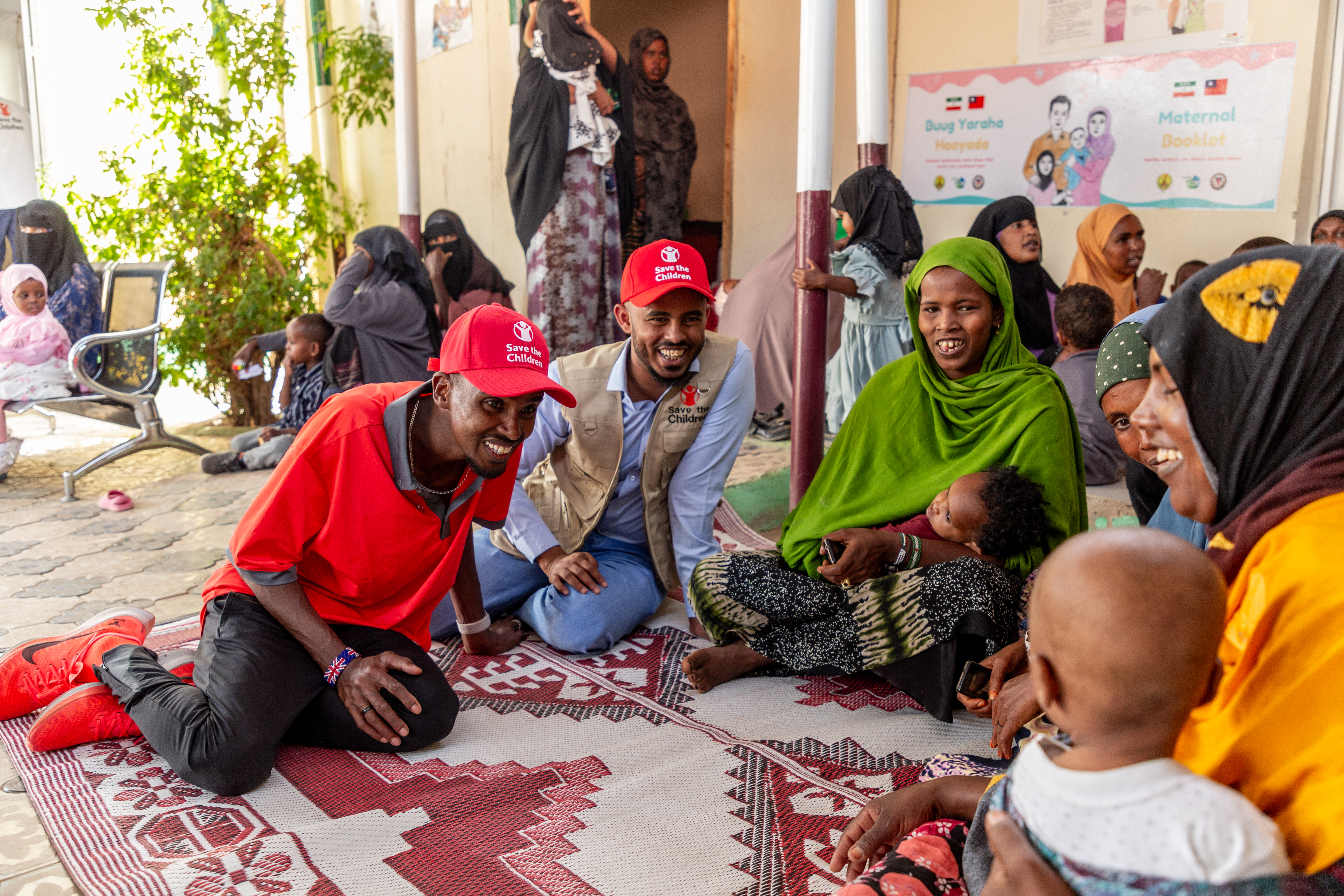 Sir Mo Farah, wearing a red Save the Children cap, sits on a rug on the floor and smiles at a mother and her young child on a visit to a health centre in Gabiley in Somaliland,