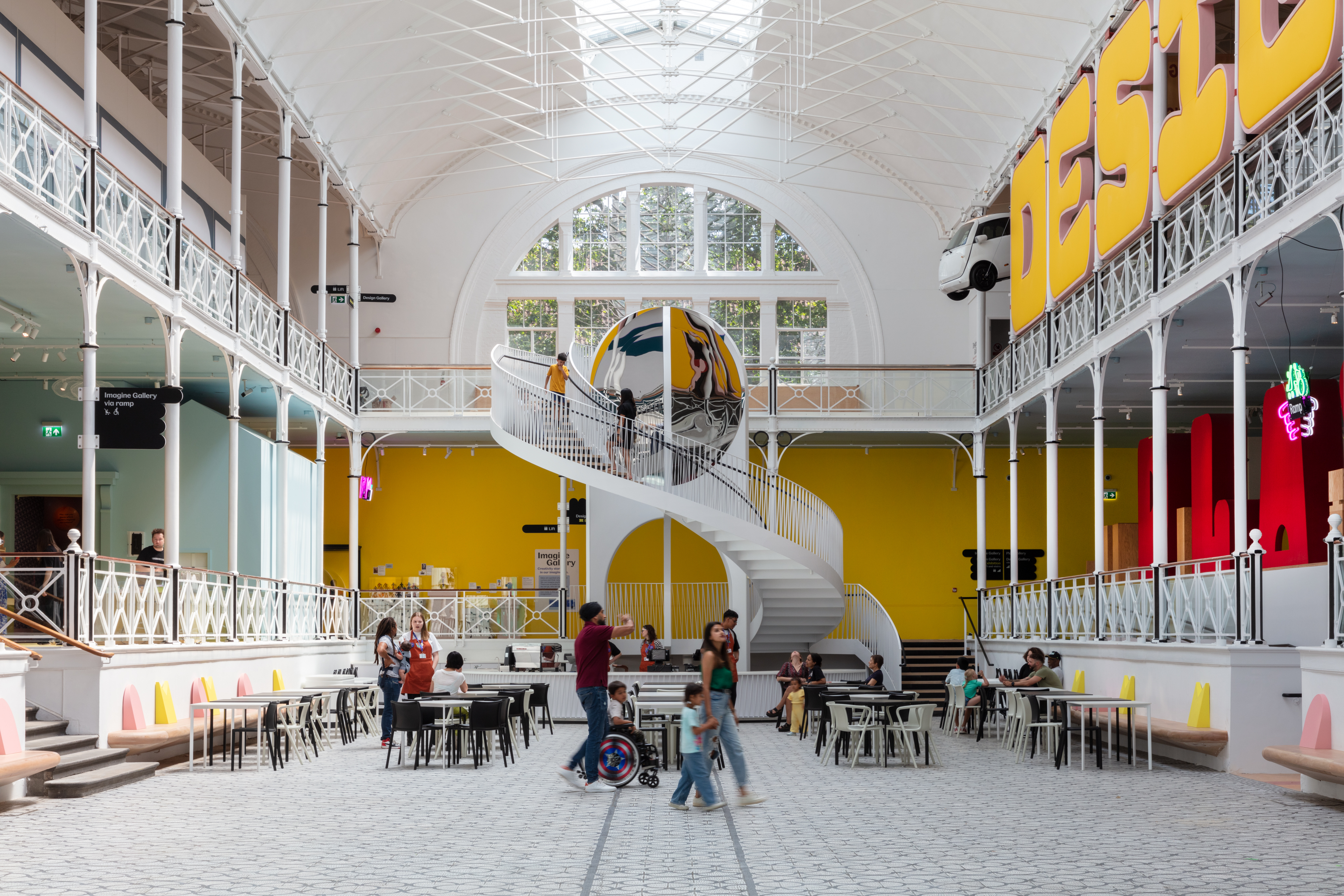 Inside the entrance of the Young V&A museum with people at tables and a large central spiral staircase leading to a mezzanine floor and window