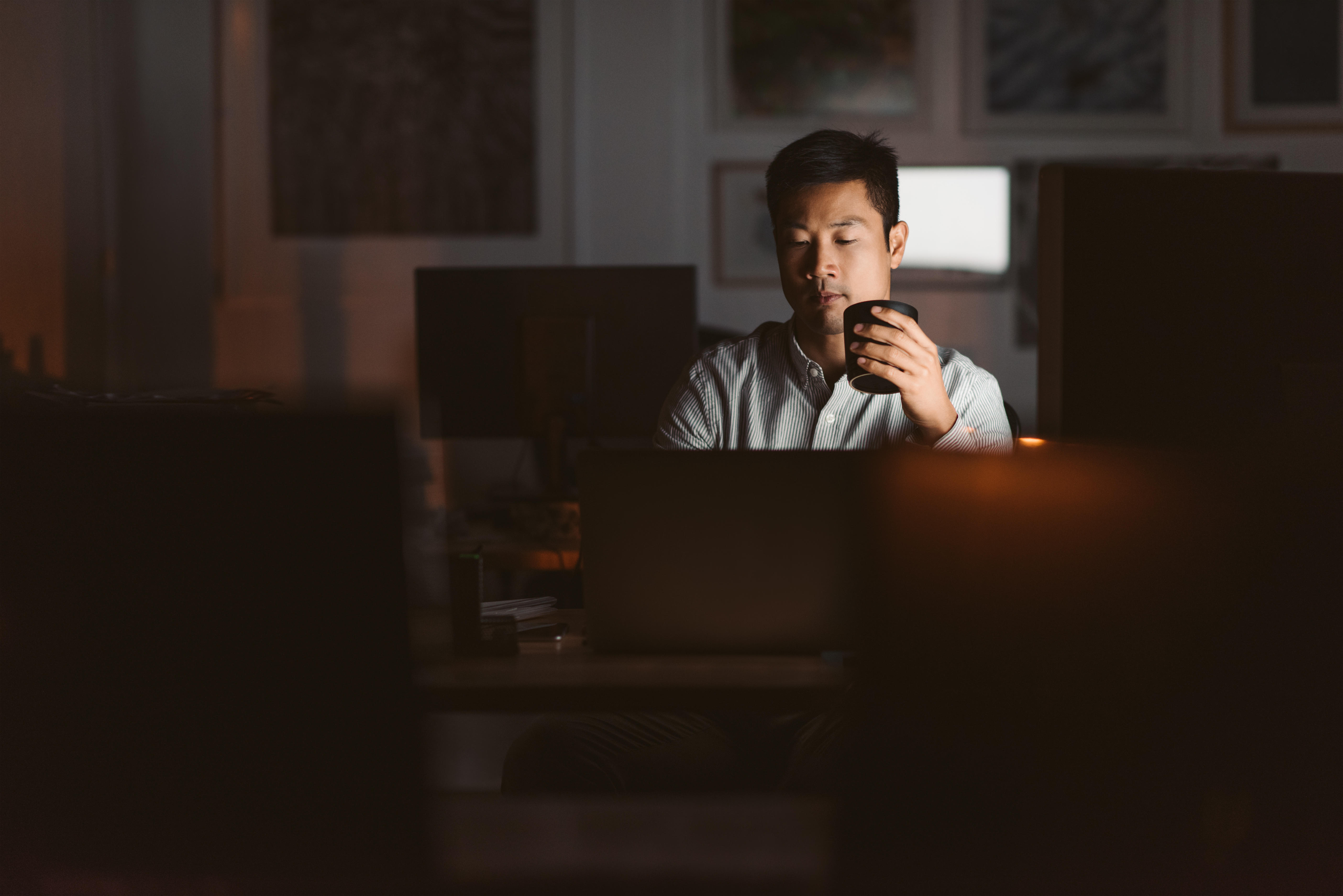 Young Asian businessman working at his desk in an office late at night drinking a cup of coffee to stay awake