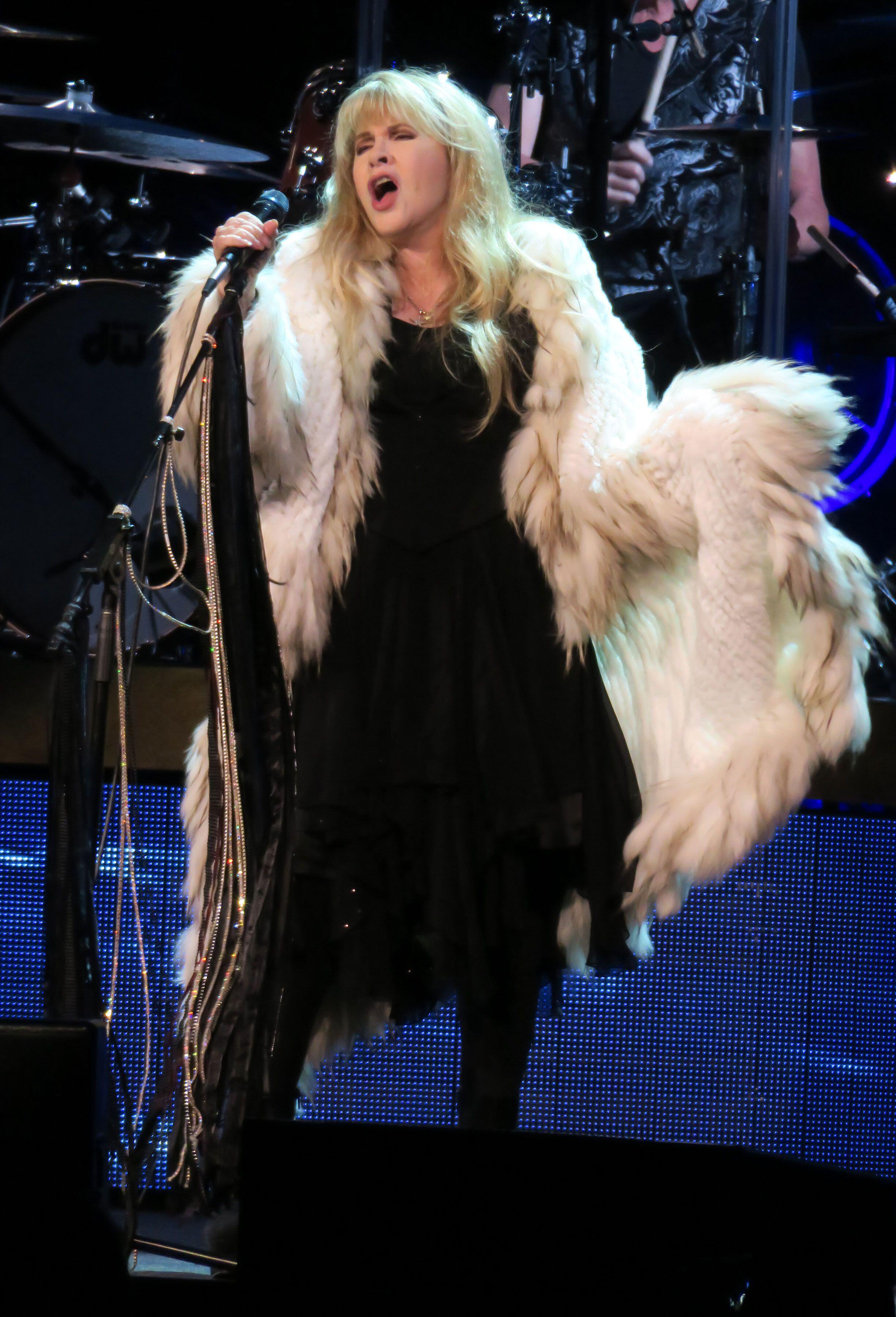 Stevie Nicks sings into mic on stage with white fur coat and black midi dress with black boots