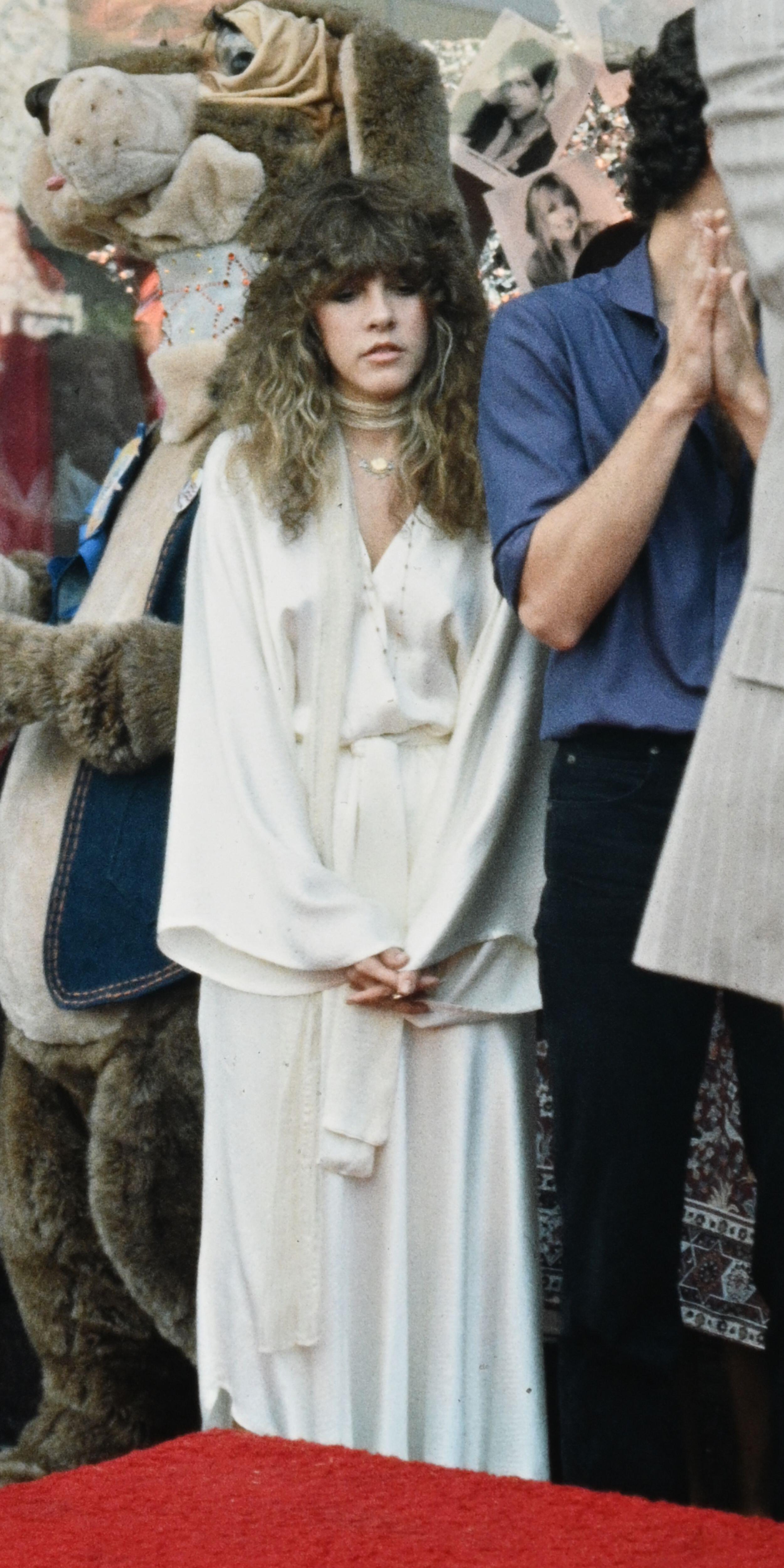 Nicks stands at the side of the stage next to bandmates in a silk cream two piece with flowing sleeves and a matching skinny scarf