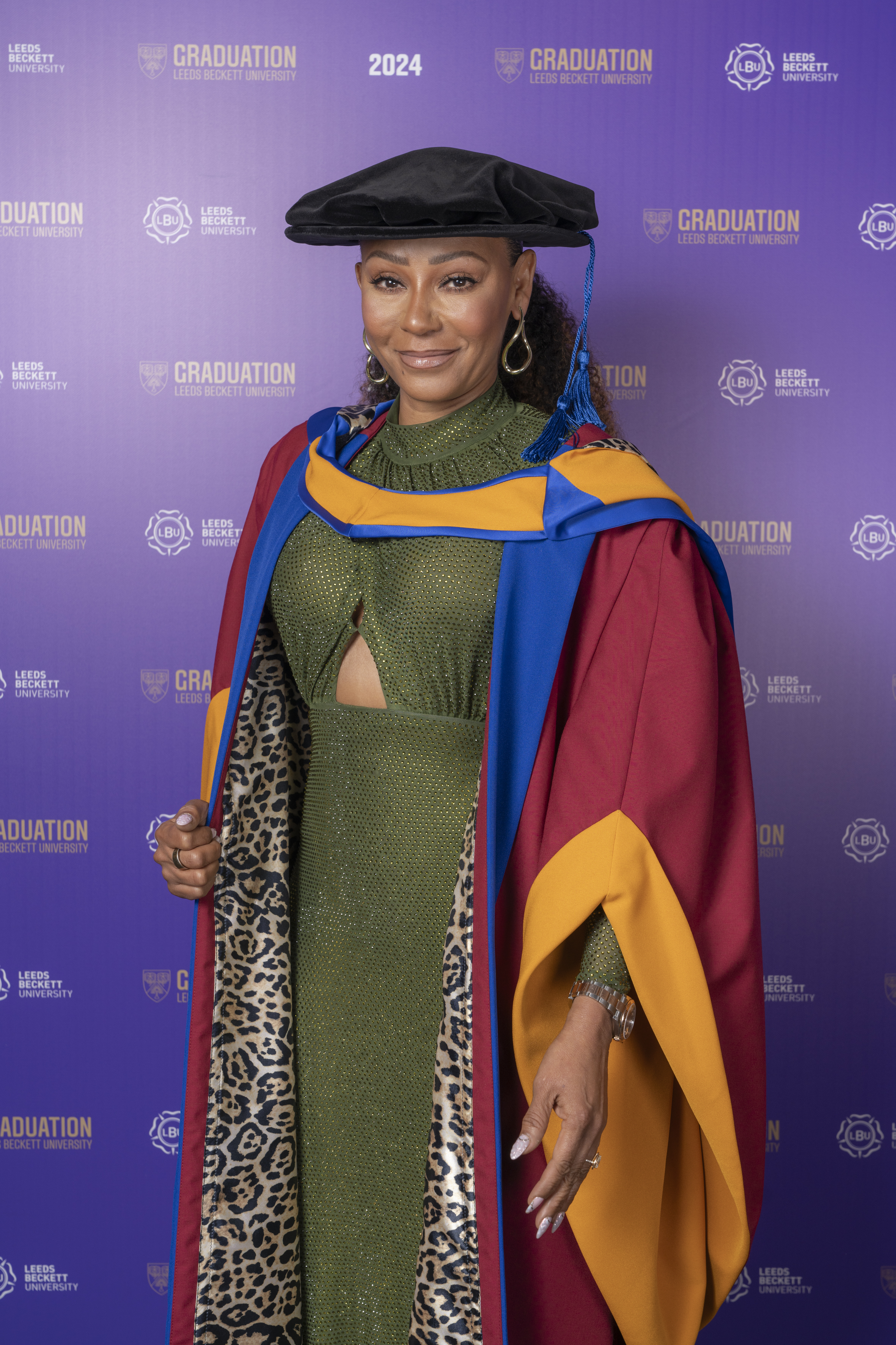 Melanie Brown in green dress and graduation gown and cap in front of purple Leeds Becket 2024 Graduation backdrop