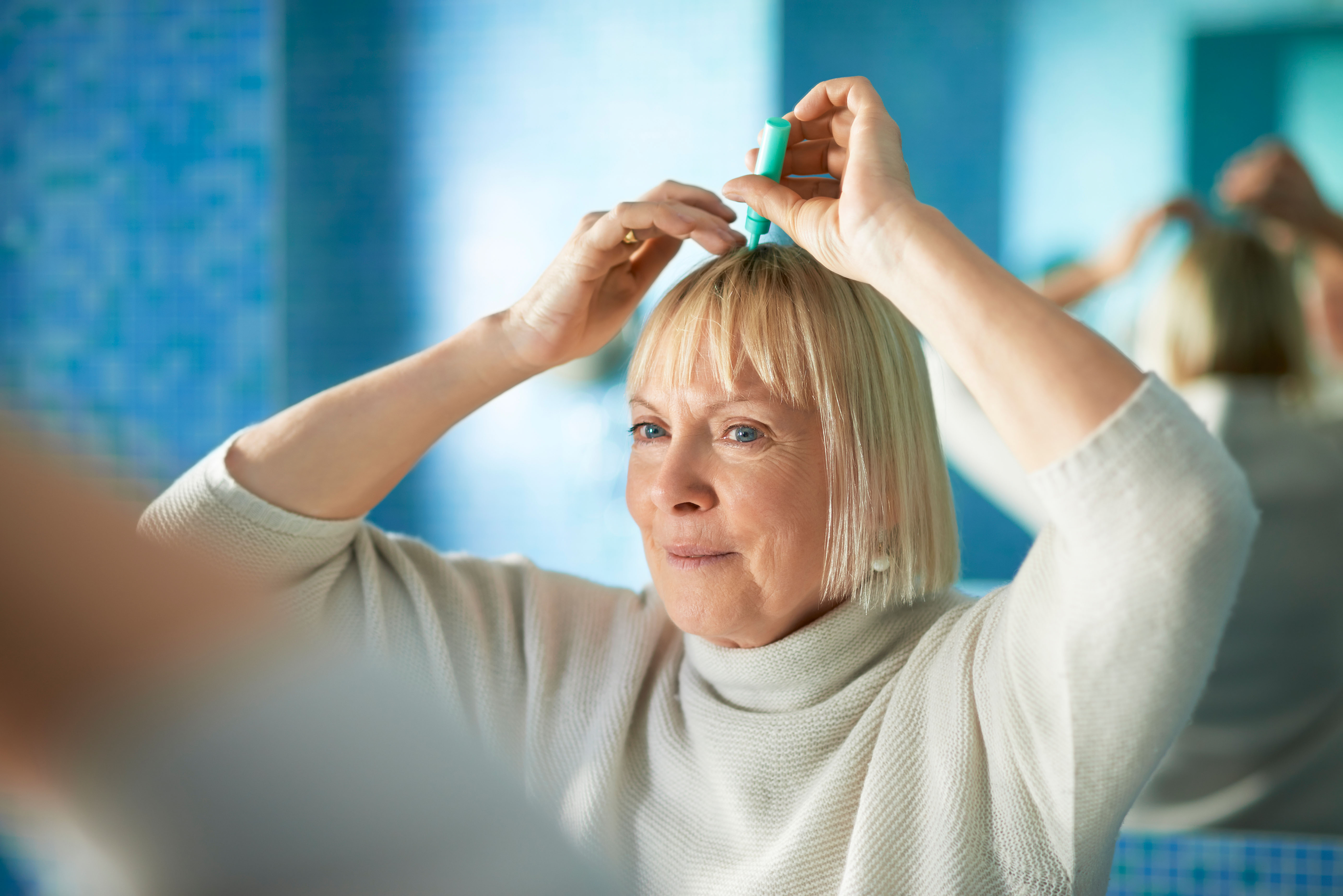 old caucasian woman applying lotion to prevent hair loss, looking at mirror in bathroom