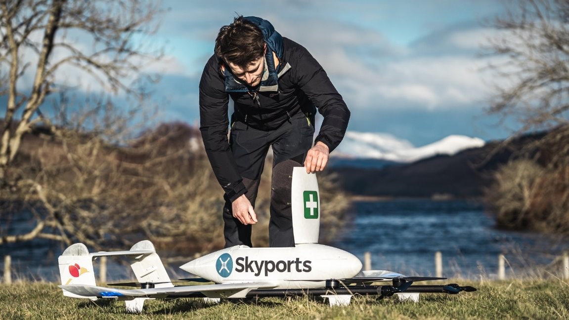 A man opens up a compartment labelled with a first aid cross on a drone, with a loch and mountains in the background