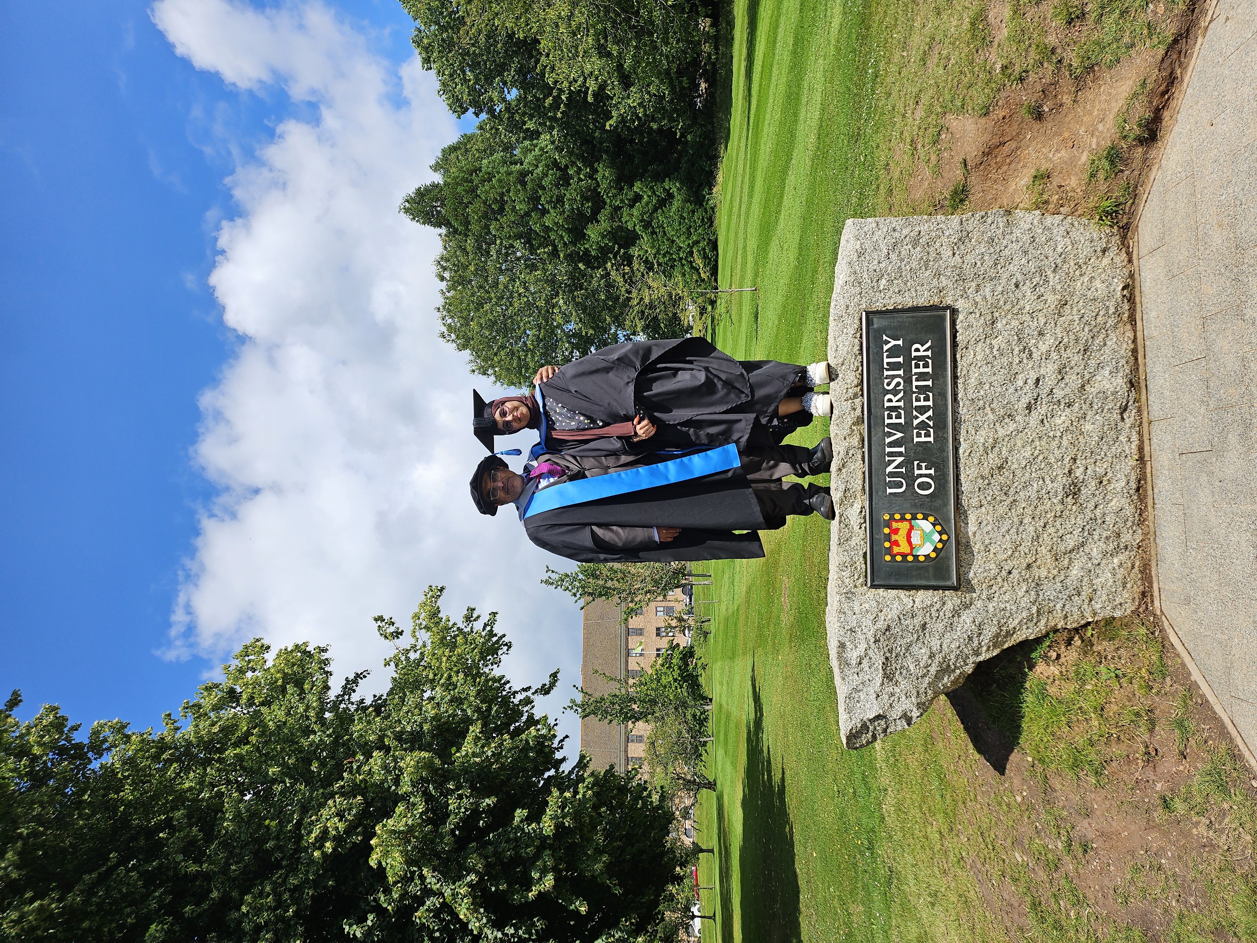 Abdallah and Alhanof Alharbi in their graduation robes and caps standing together on a stone sign for the University of Exeter