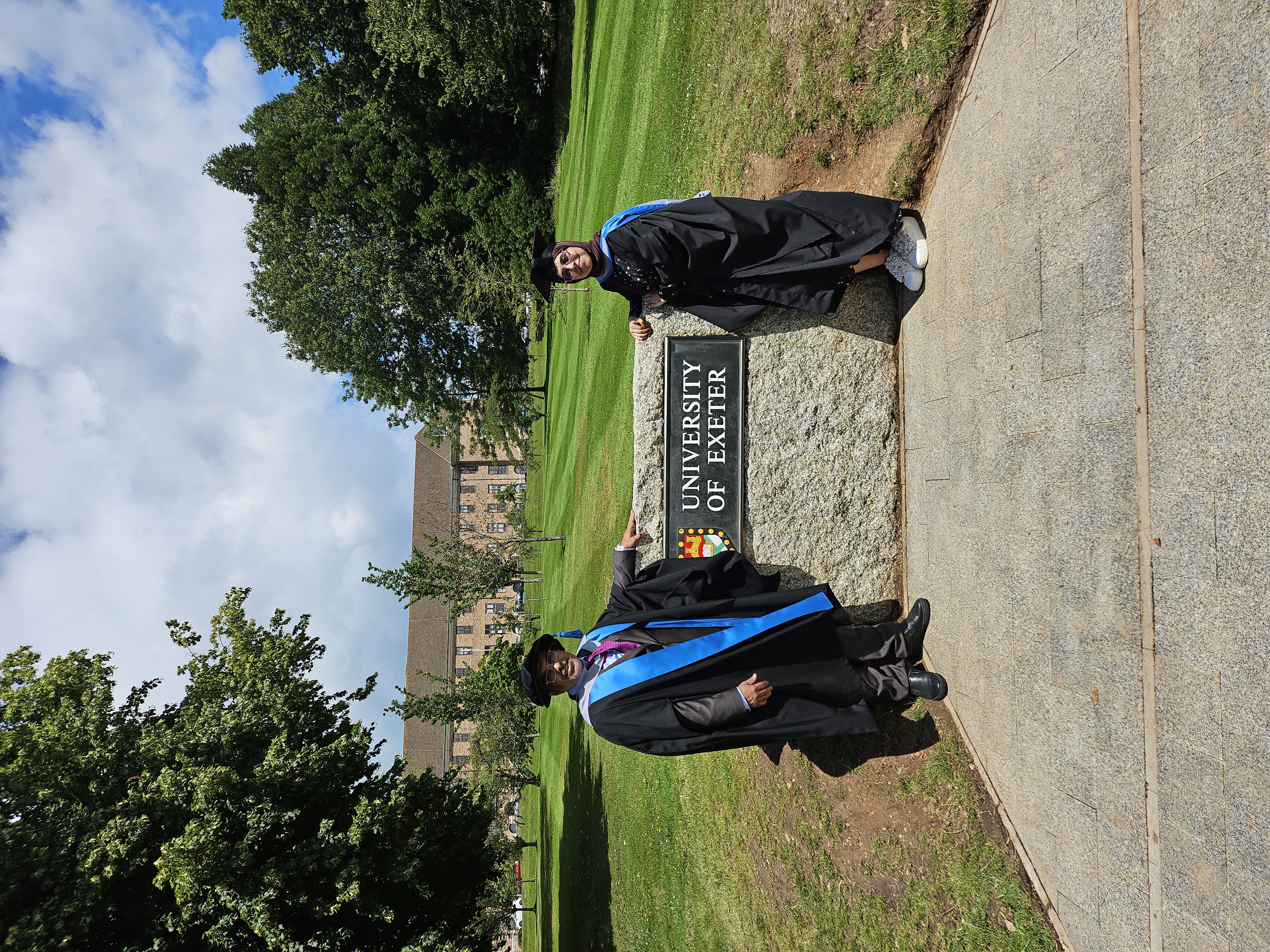 Abdallah and Alhanof Alharbi either side of a University of Exeter sign surrounded by trees and lawn