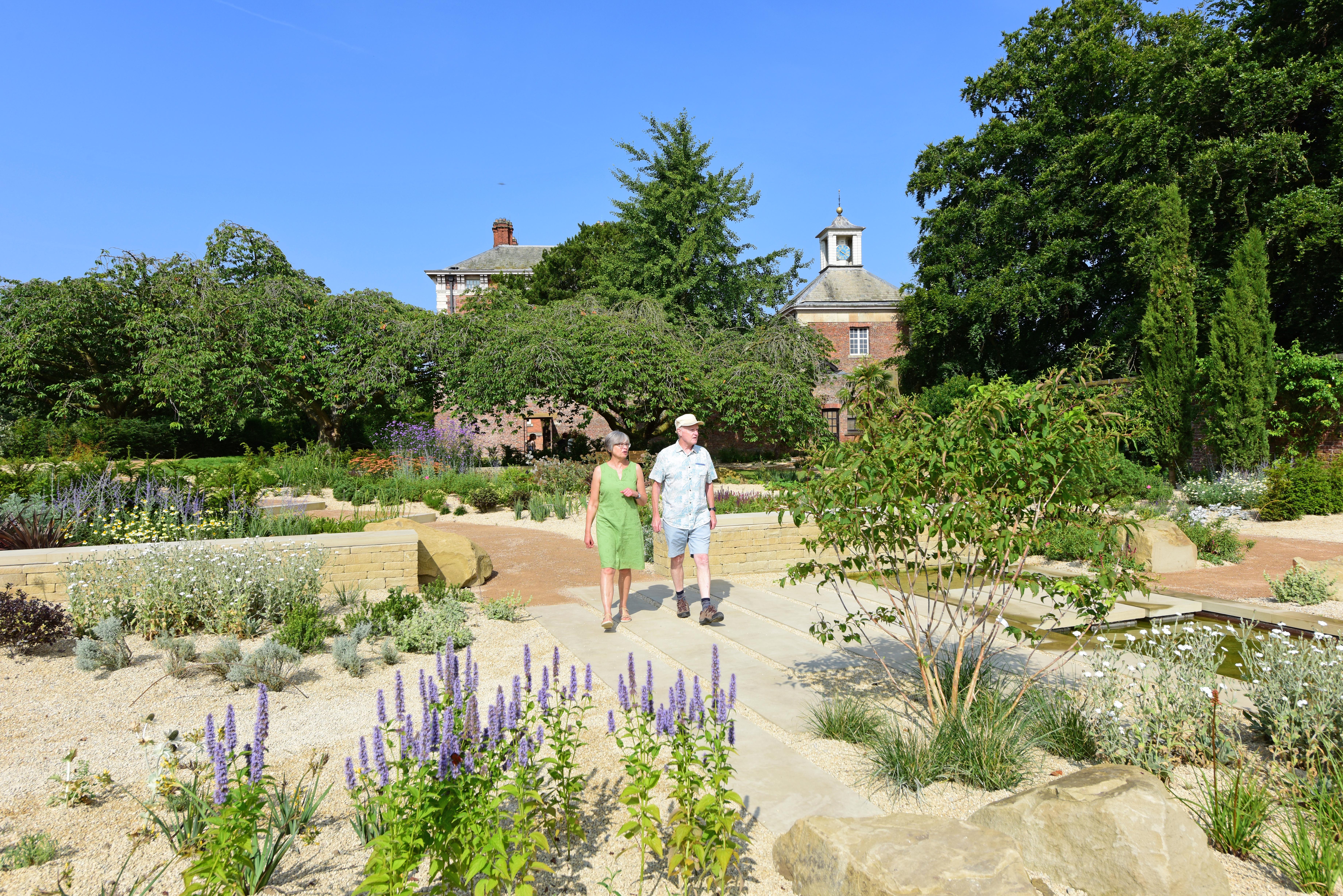 A couple walk through the garden, with gravel, flowers, young trees in the foreground, and trees and parts of Beningbrough Hall behind