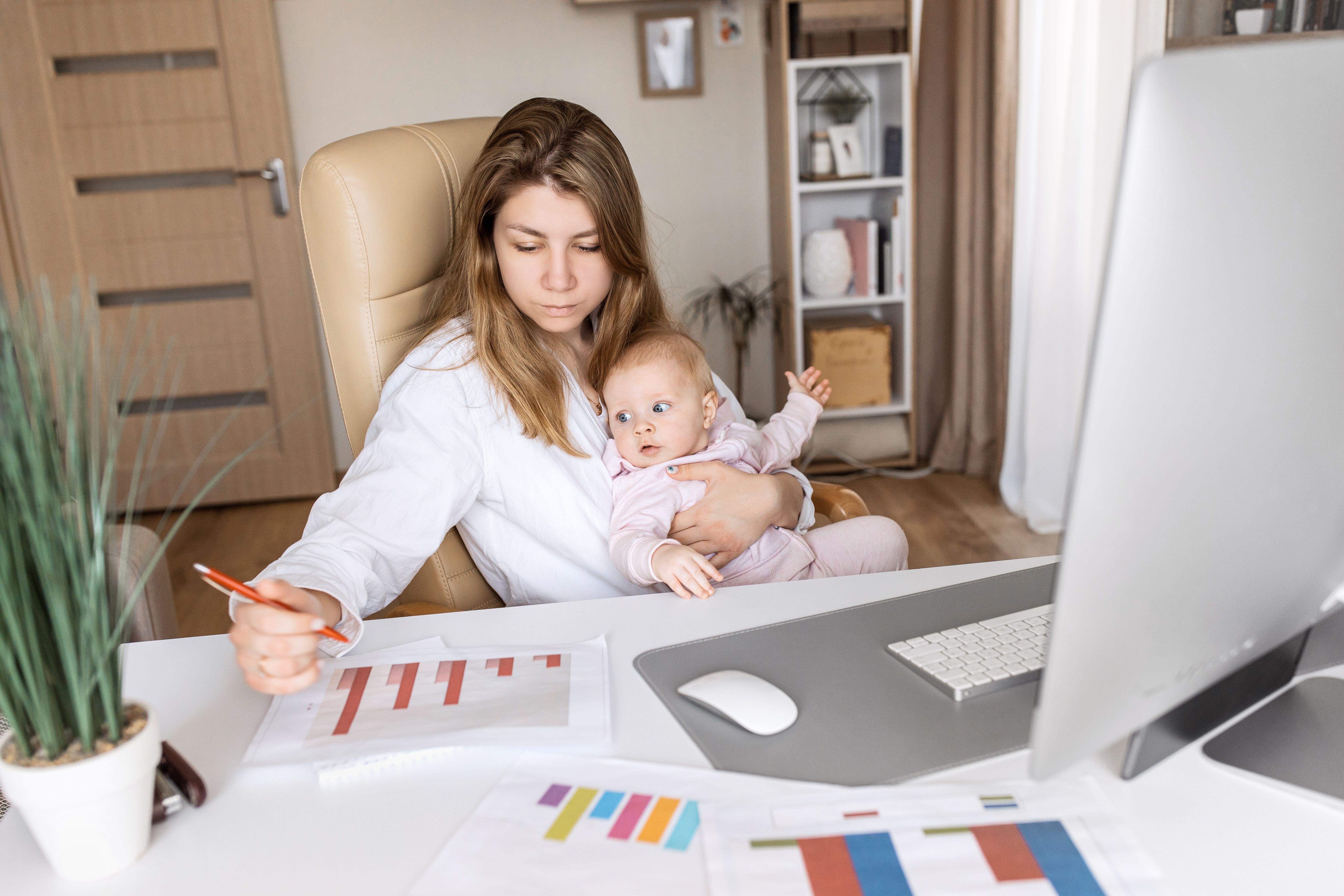 A concerned woman sitting at a desk looking at bar charts while also holding a baby