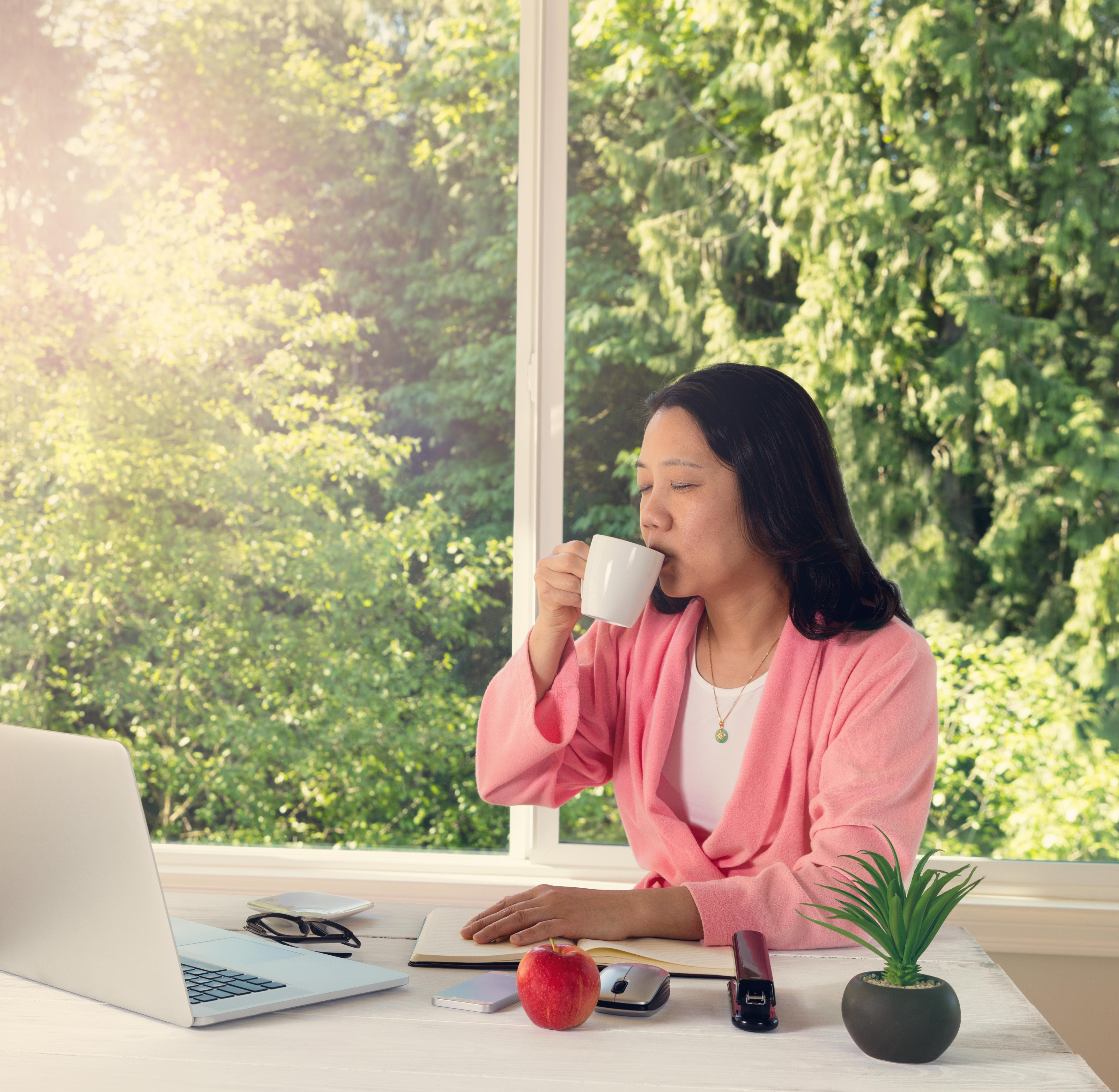 A woman enjoying her morning coffee while working from home in front of large daylight window