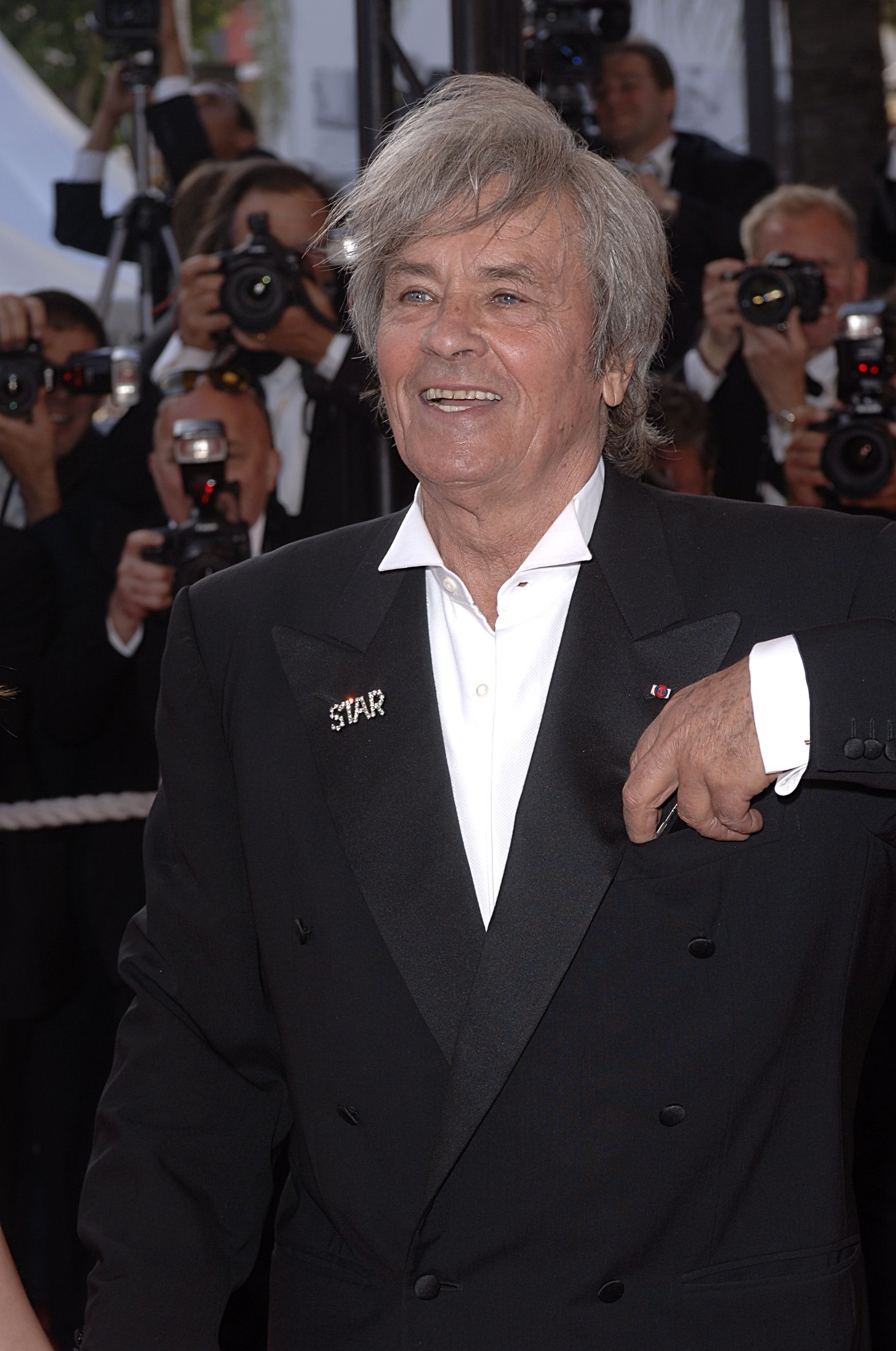 Alain Delon smiling in suit jacket, in front of a bank of photographers