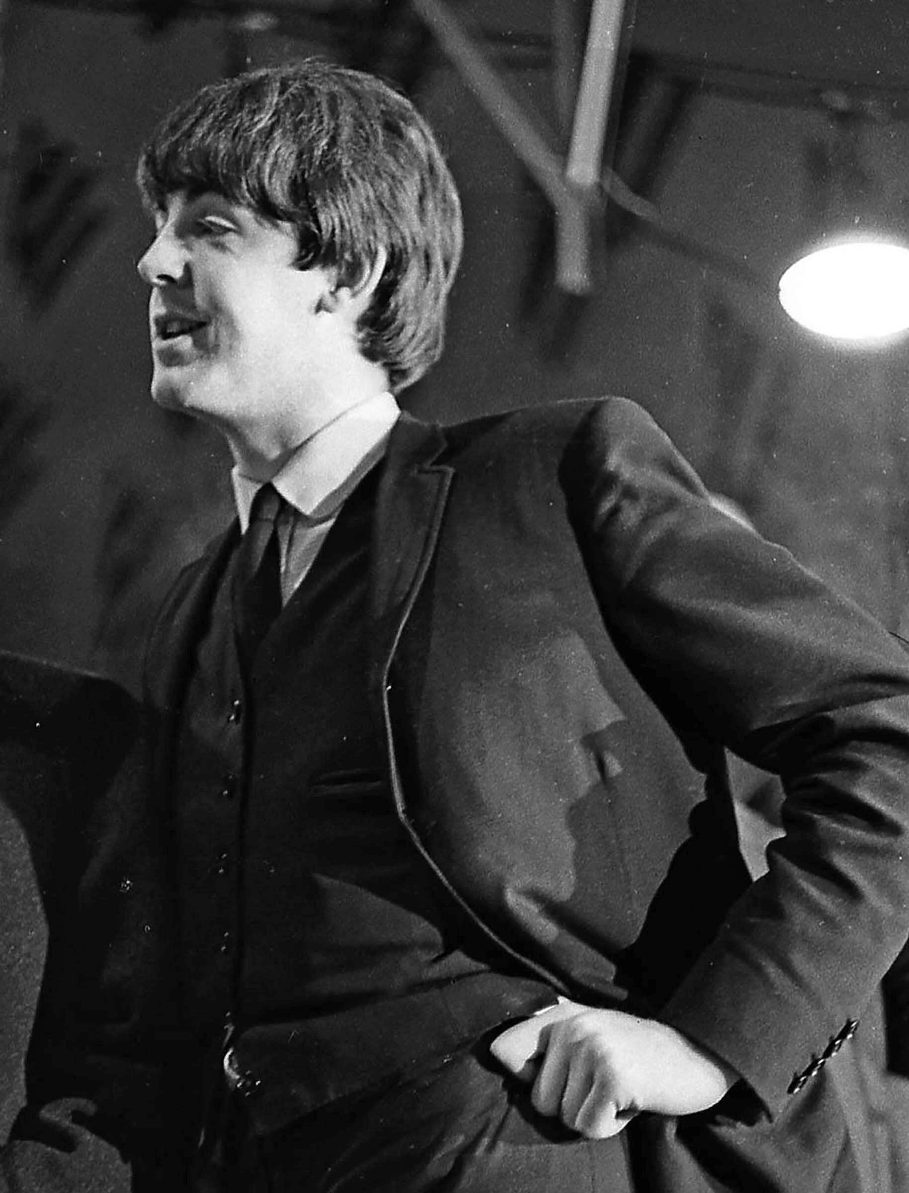 Paul McCartney stands on stage in black white and tie with white shirt