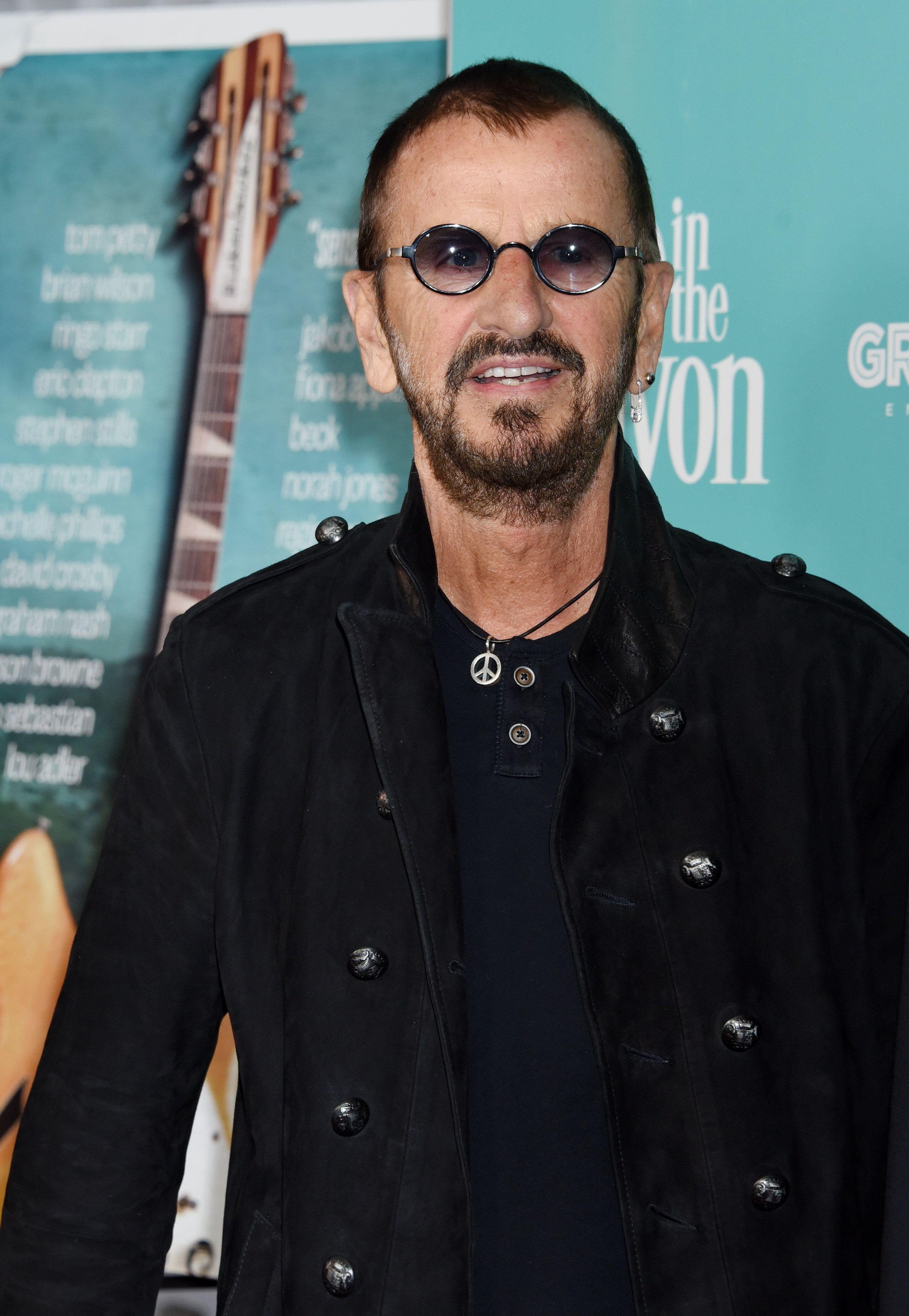 Ringo Starr wears black T-shirt and jacket and round wire rimmed glasses with silver peace necklace