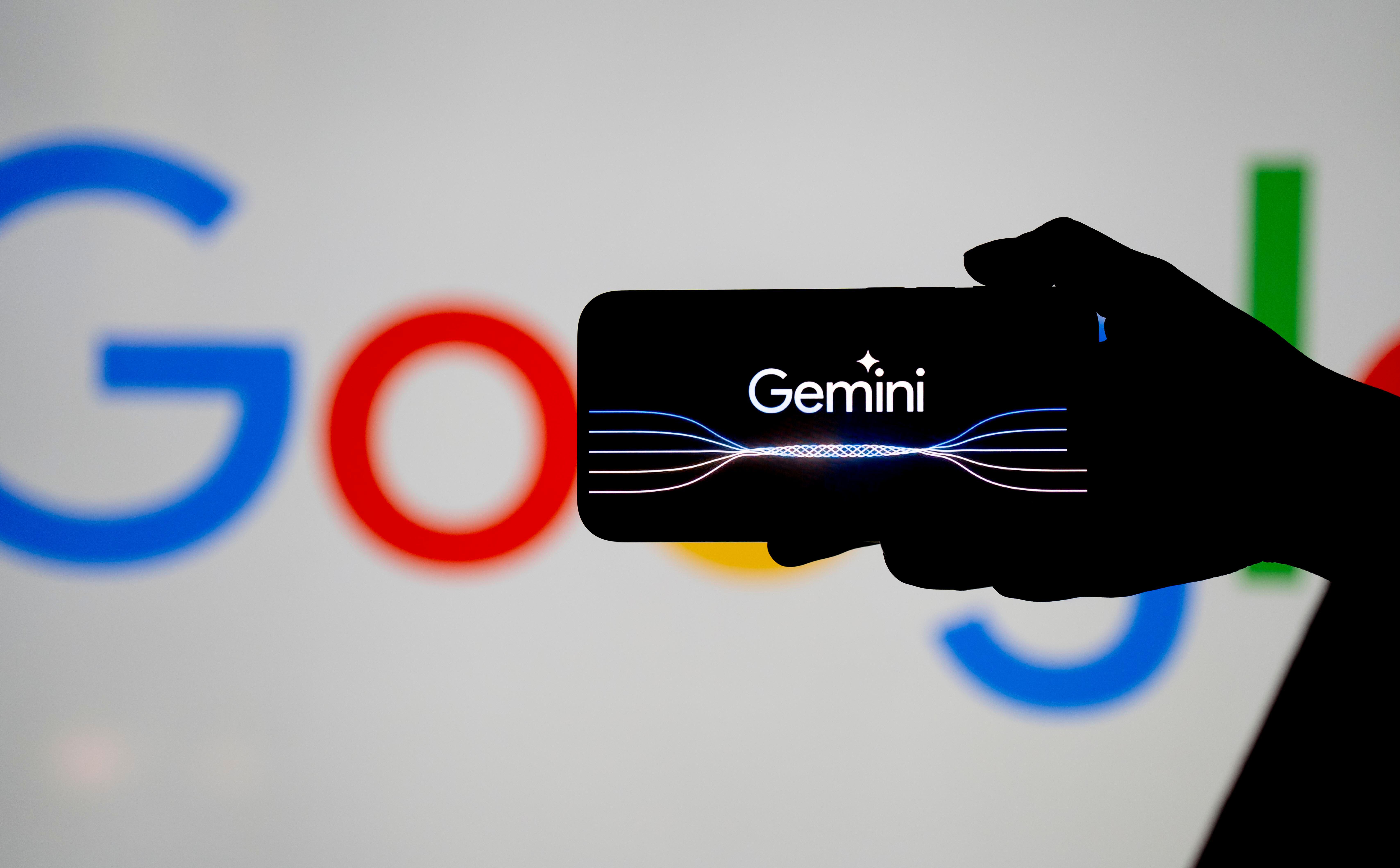 Silhouette of a hand holding a phone with the Gemini logo on its screen with the Google logo partially obscured in the background