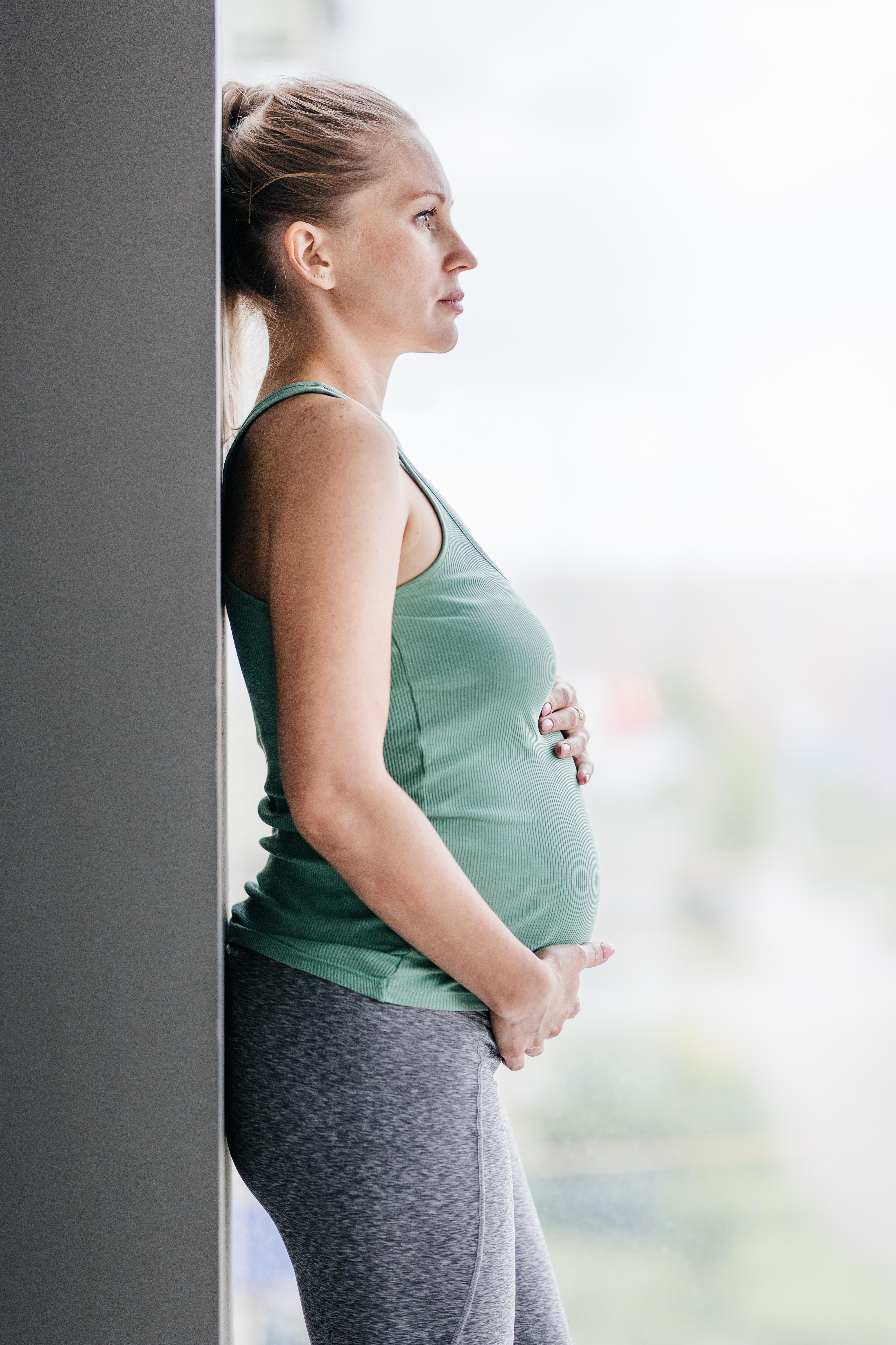 Pregnant woman holding her belly standing near a window wearing sportswear 