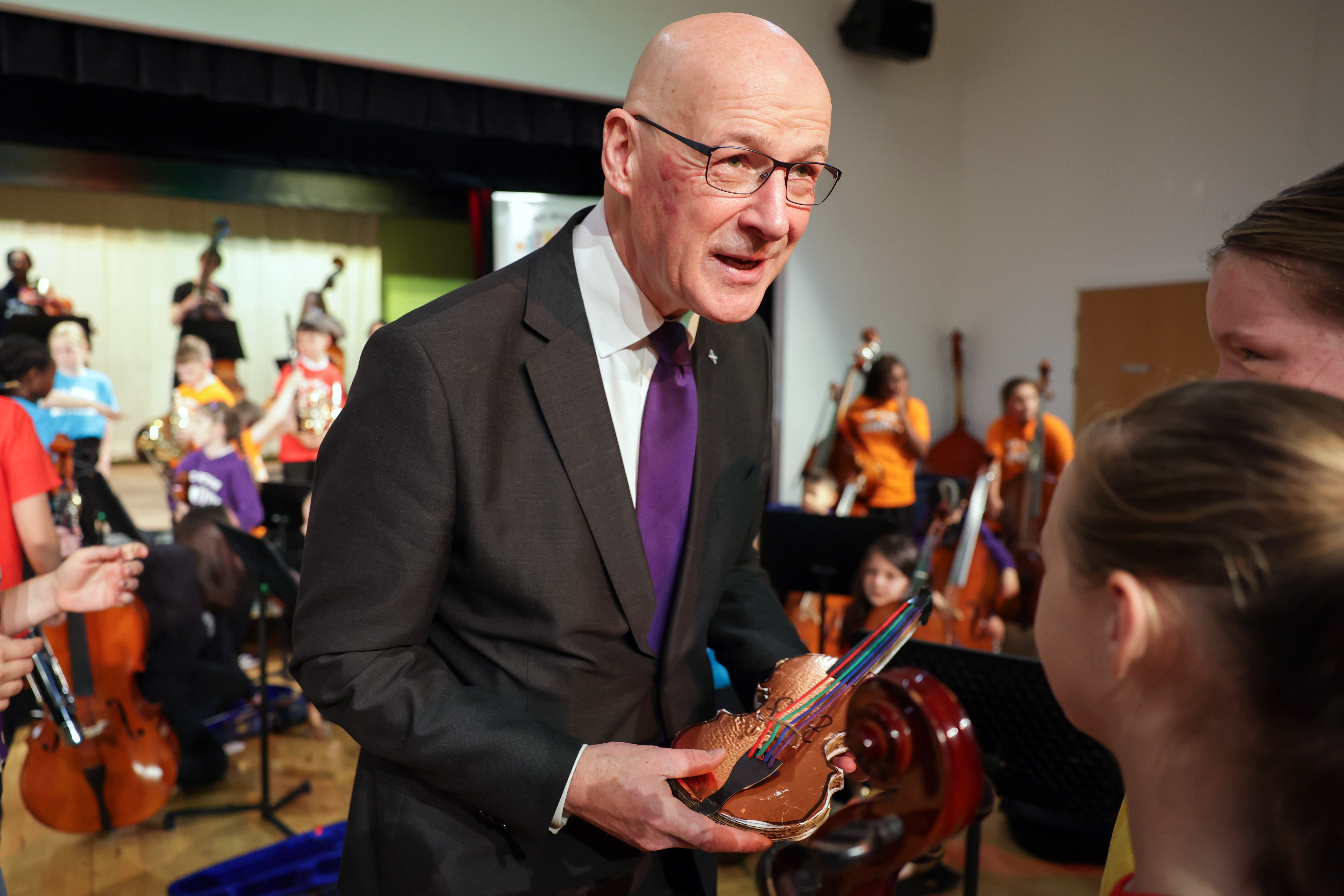 John Swinney holding a violin, while speaking to children in front of a children's orchestra