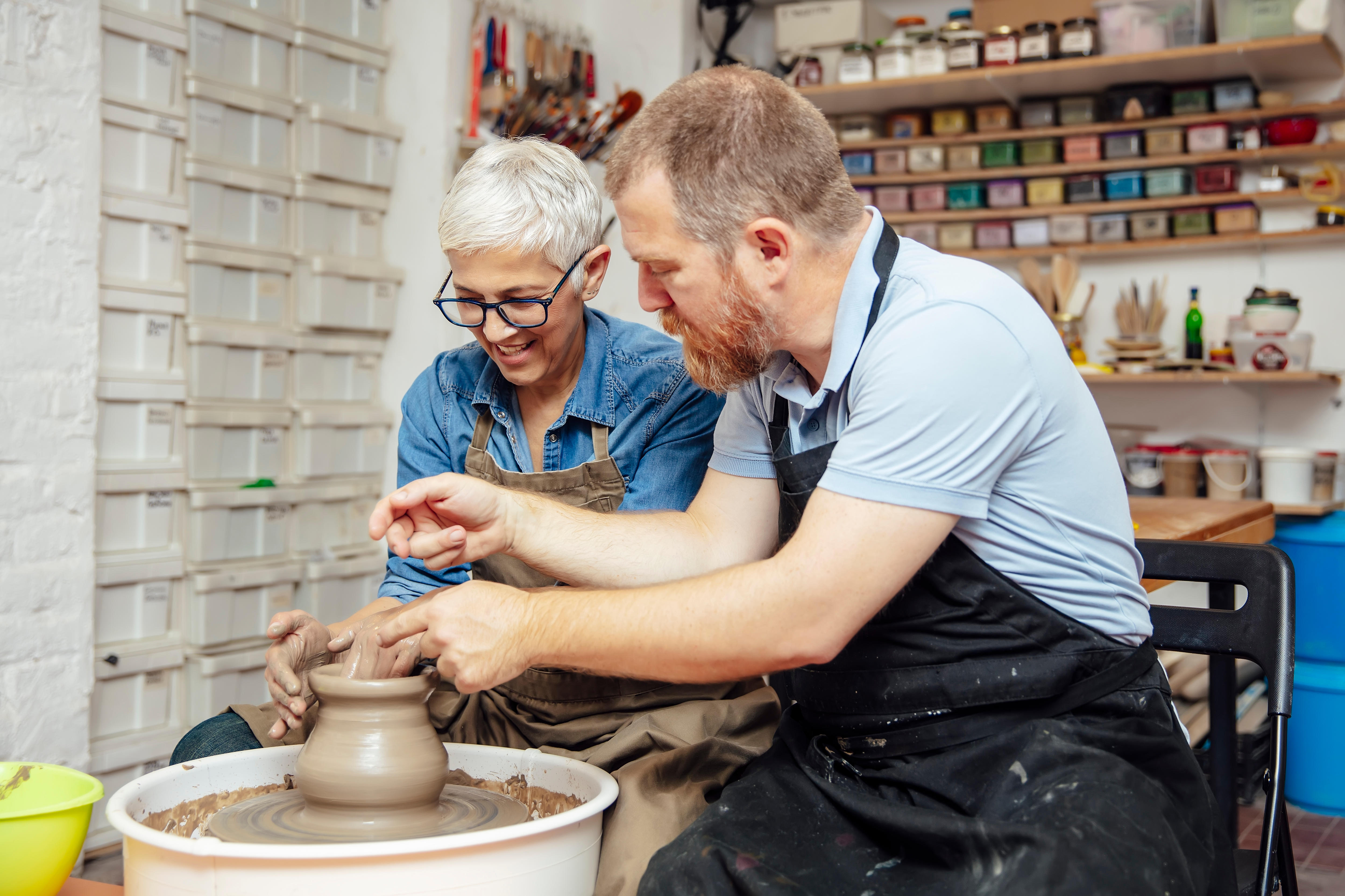 Senior woman spinning clay on a wheel with a help of a teacher at pottery class 