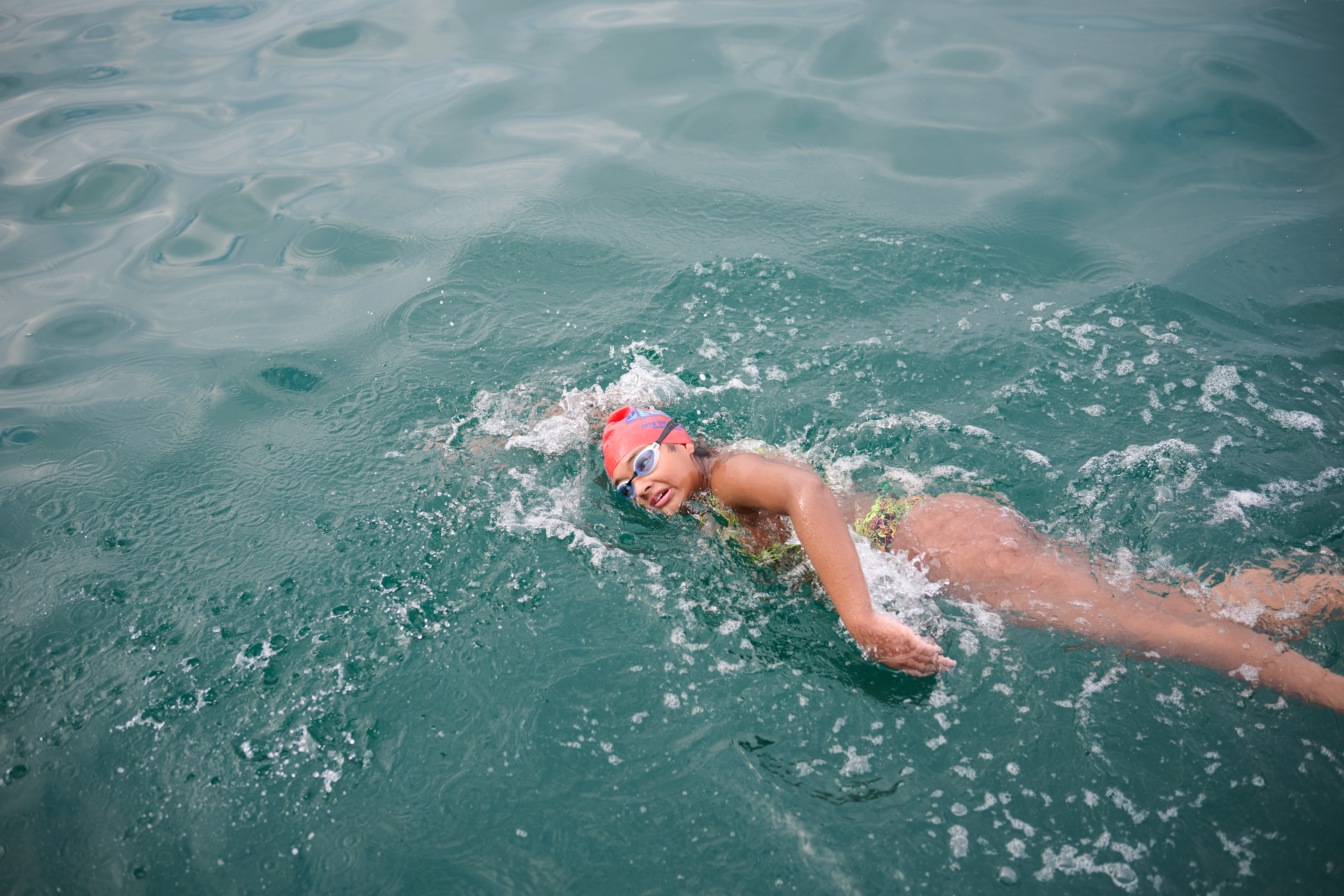 Prisha wearing a swimming cap and goggles turns her head to one side while her left arm emerges from the water as she does the front crawl