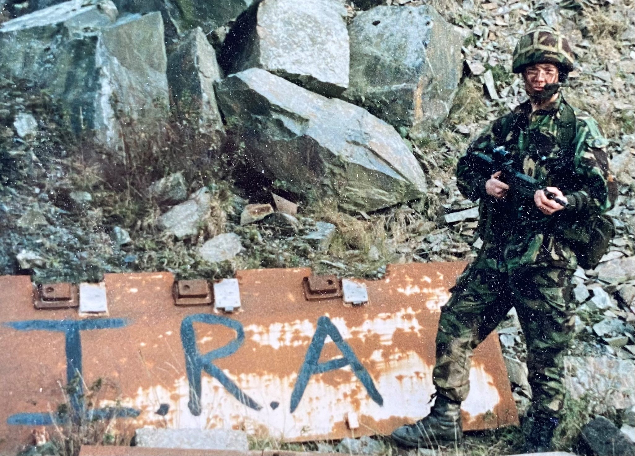 Rob Hughes in Army uniform and gun, beside graffiti reading 'IRA'