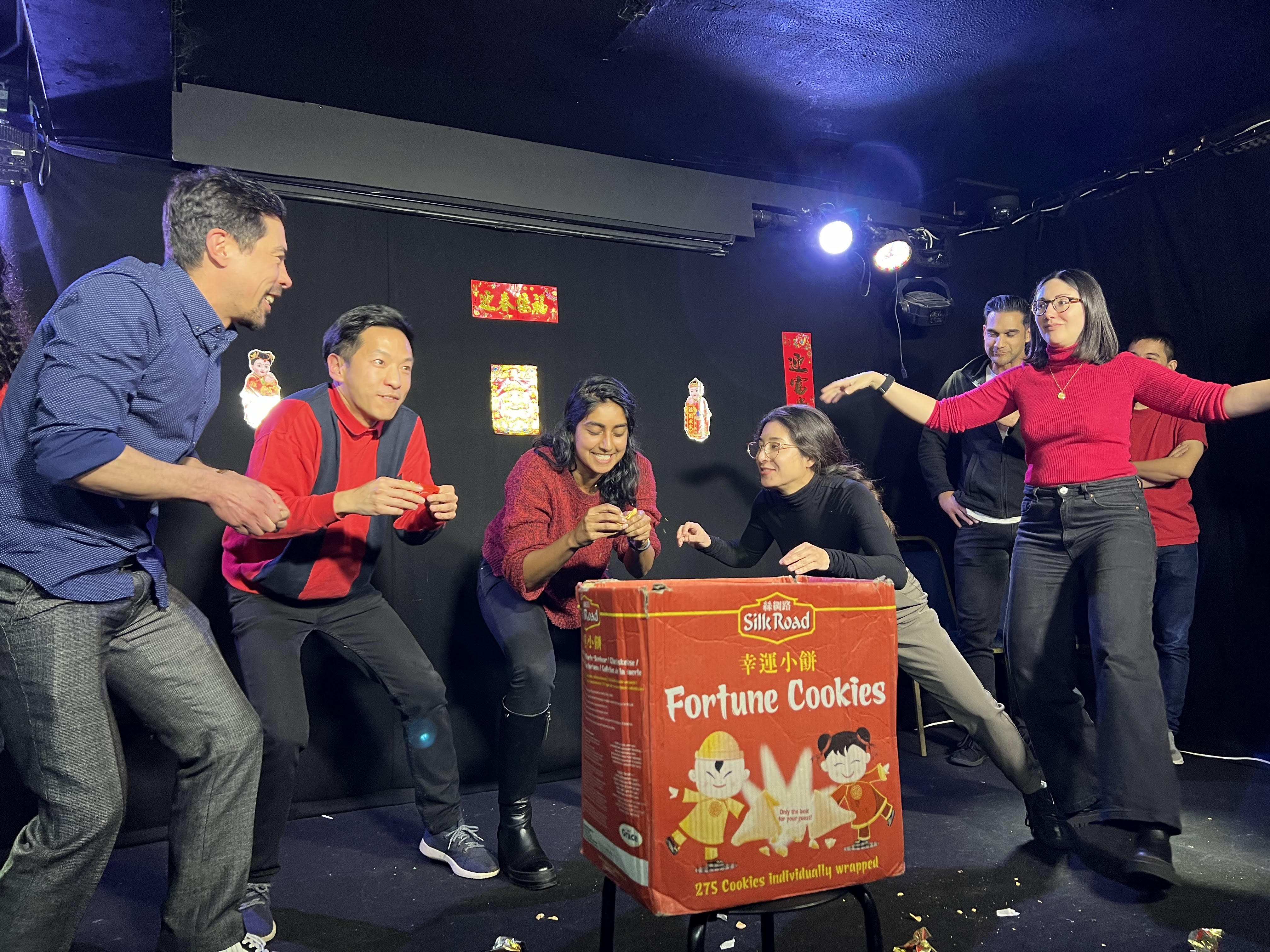 Group of people surround a red box which says fortune cookies as they perform improv on stage