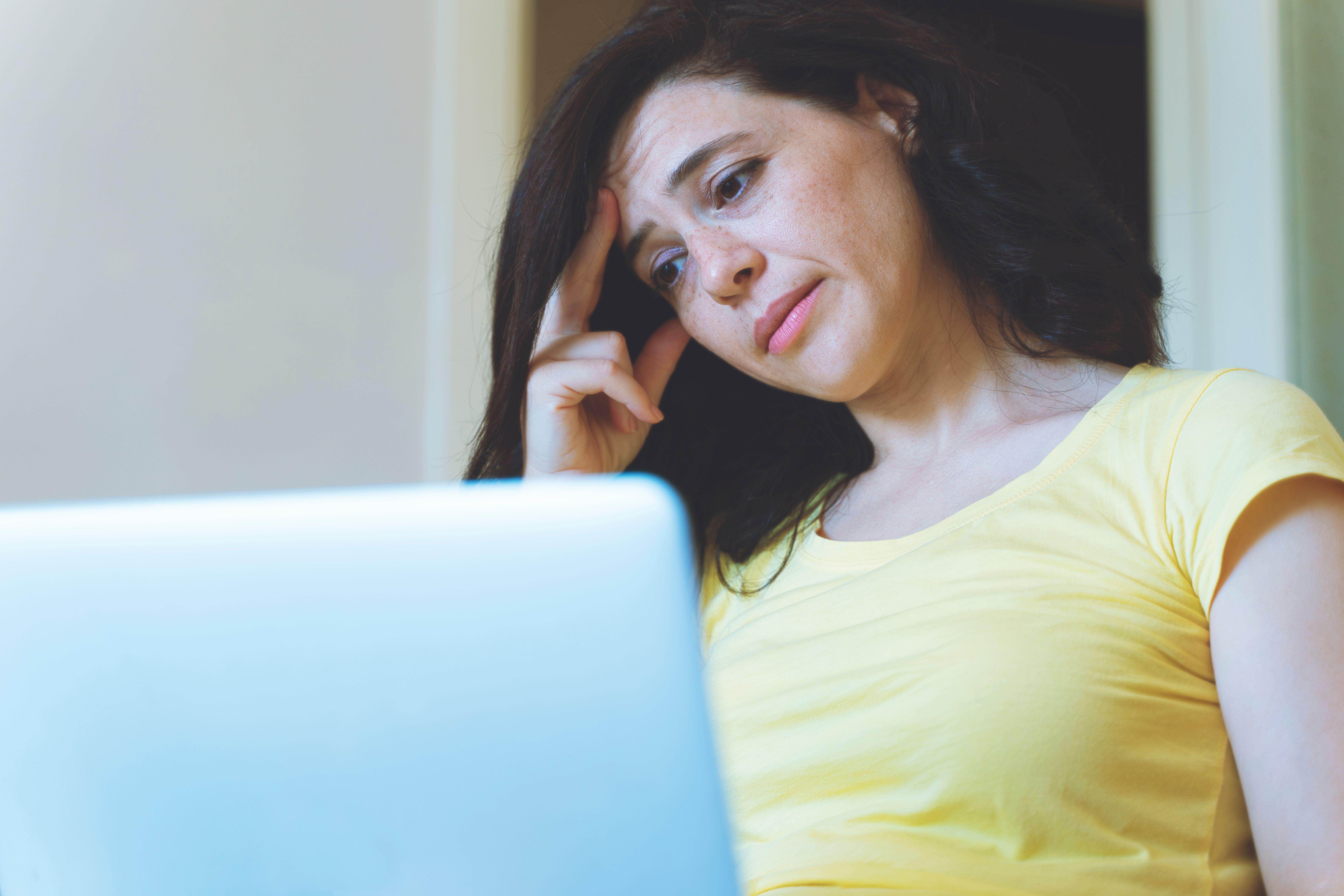 A 40-something woman in a yellow top, sitting at home with a laptop open in front of her. She looks slightly overwhelmed or anxious 