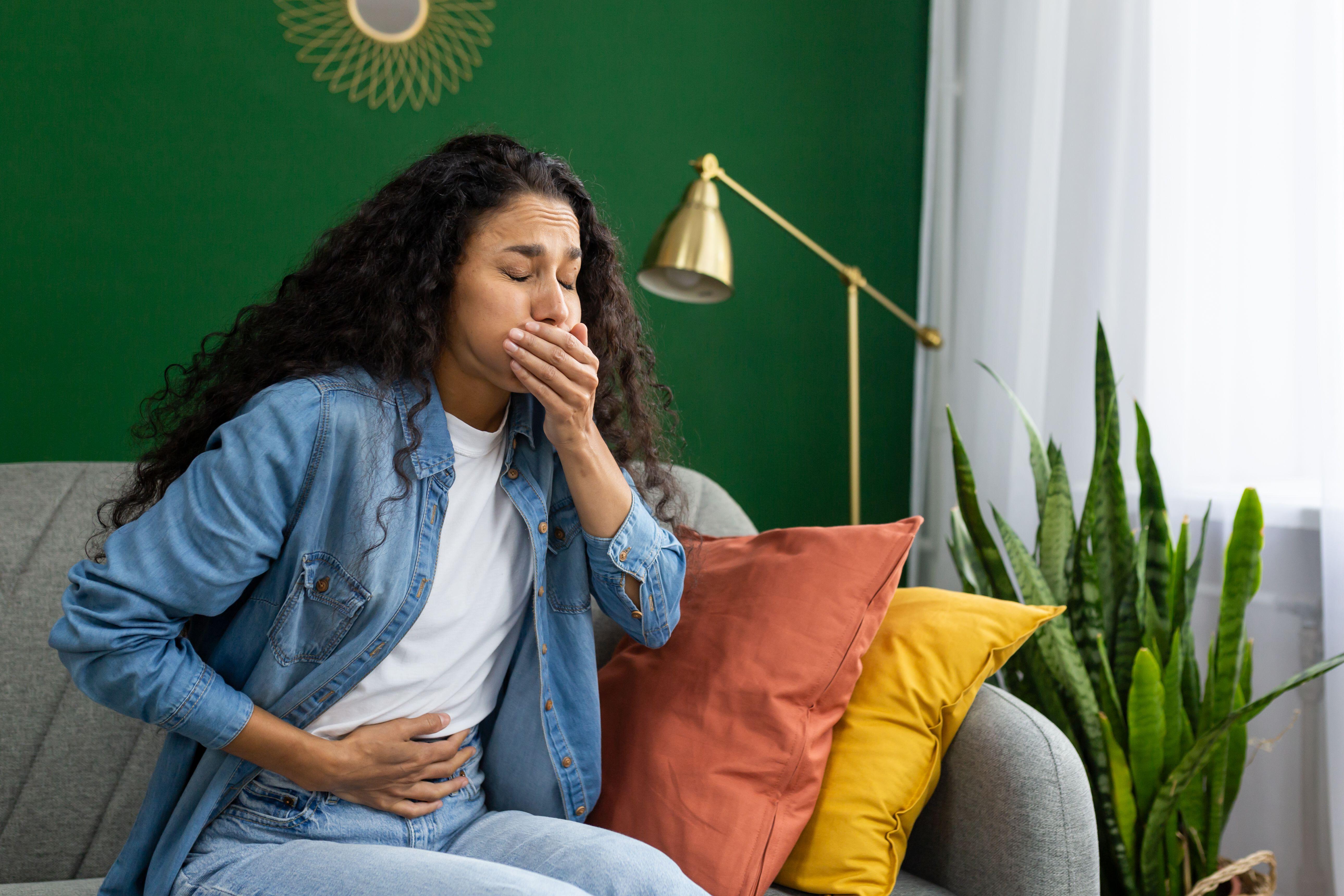 A young woman feeling nauseous on a couch, with one hand over her mouth and the other on her stomach 