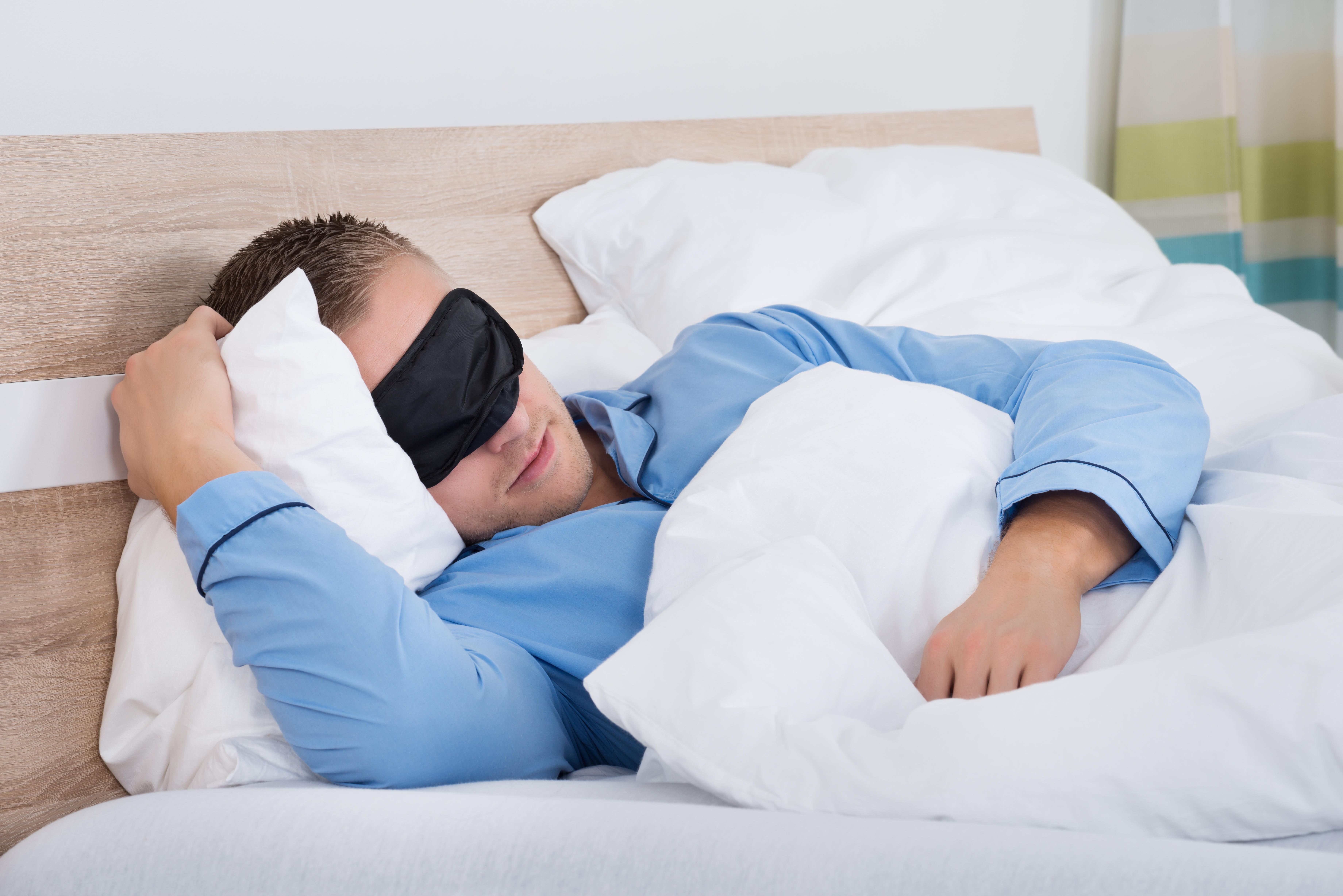 Young man wearing a blue shirt and a black eye mask sleeping in bed