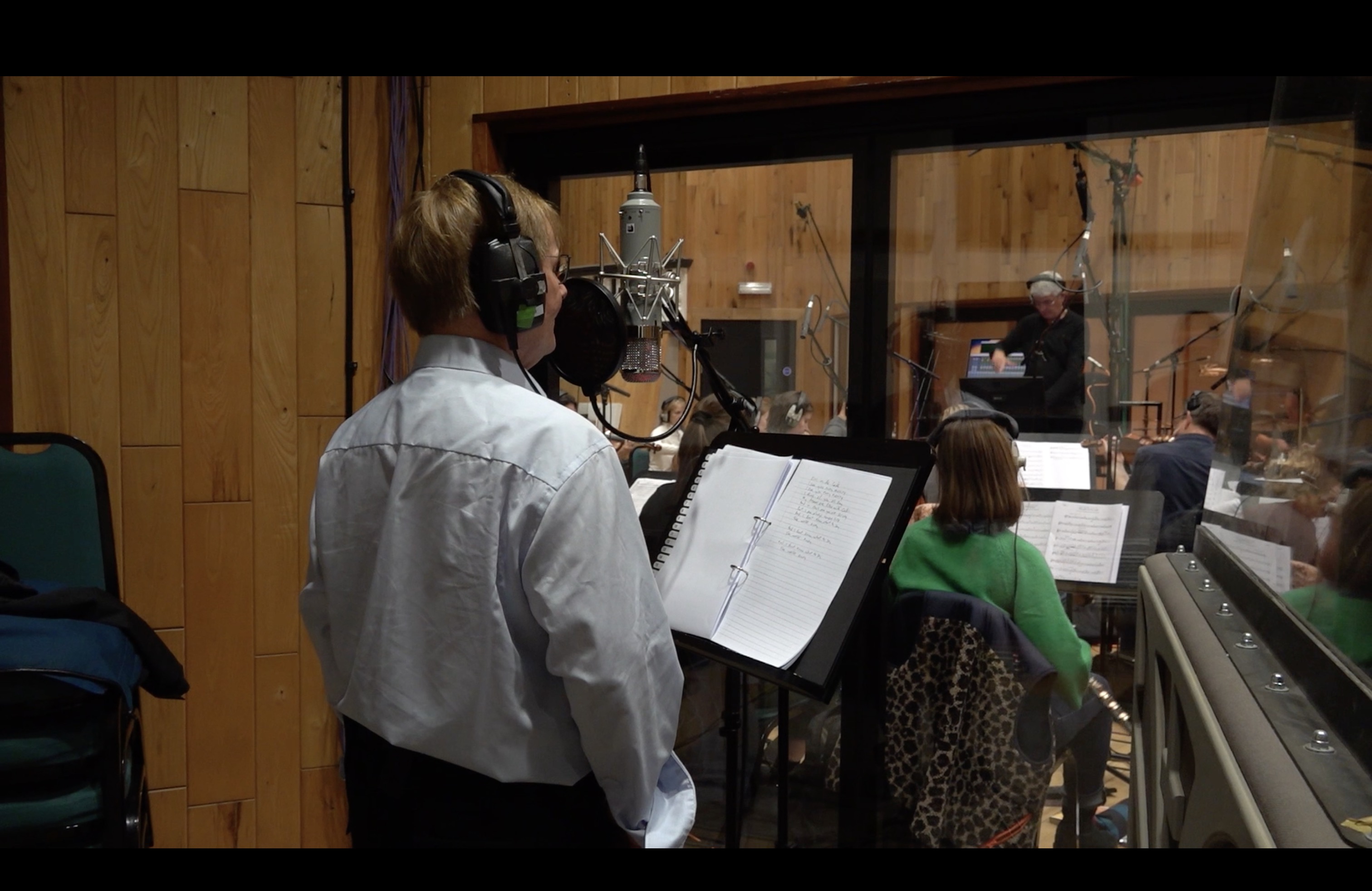 Man standing in a recording studio while an orchestra plays in front of him