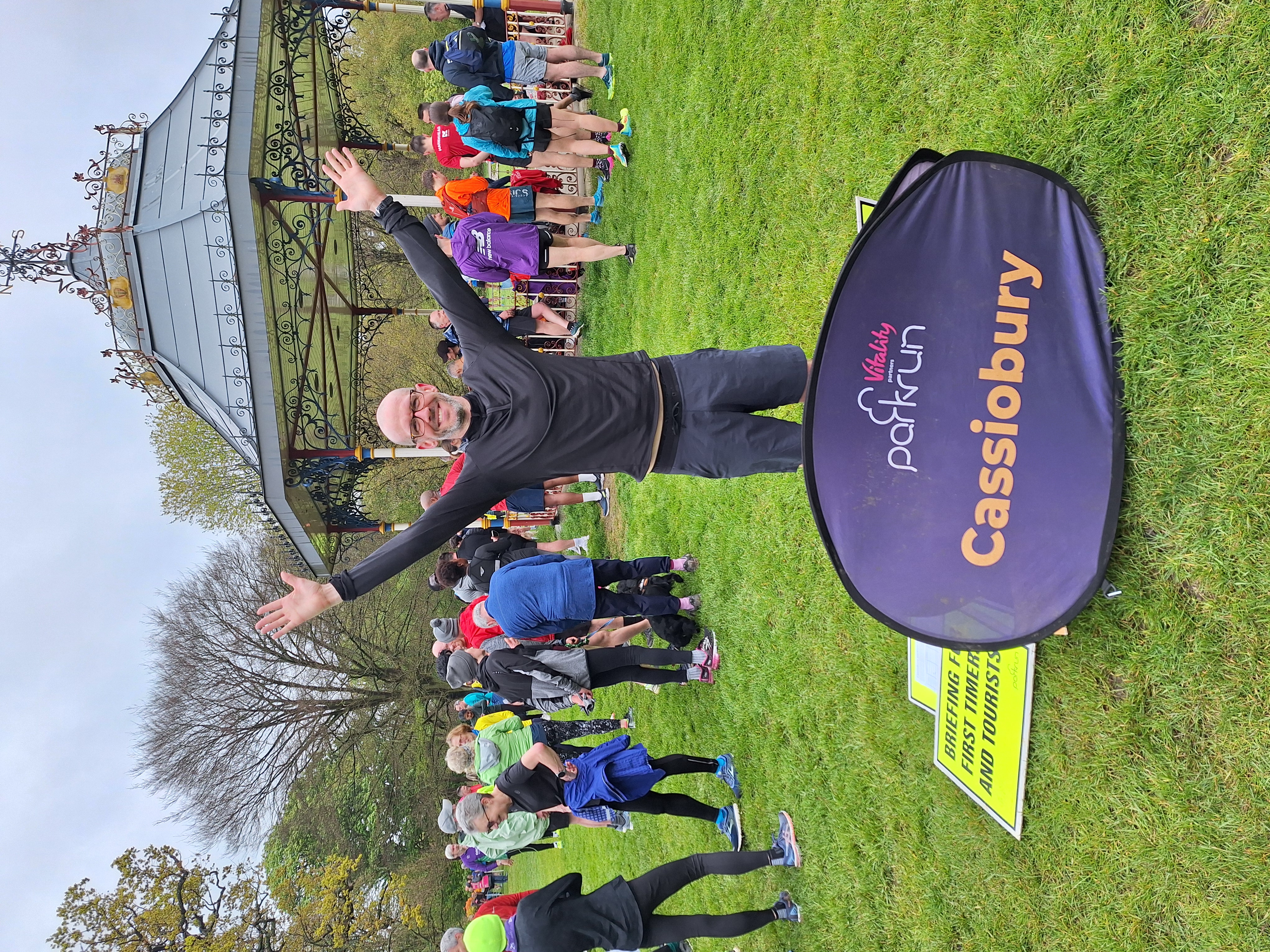 Patrick Fox with his arms in the air and the sign for Cassiobury parkrun at his 100th parkrun in April 2024