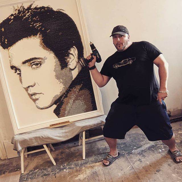 A man posing with a drill next to an artwork of Elvis