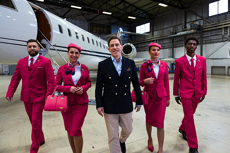 Still from Destination X showing host Rob Brydon next to plane with four people dressed as cabin crew