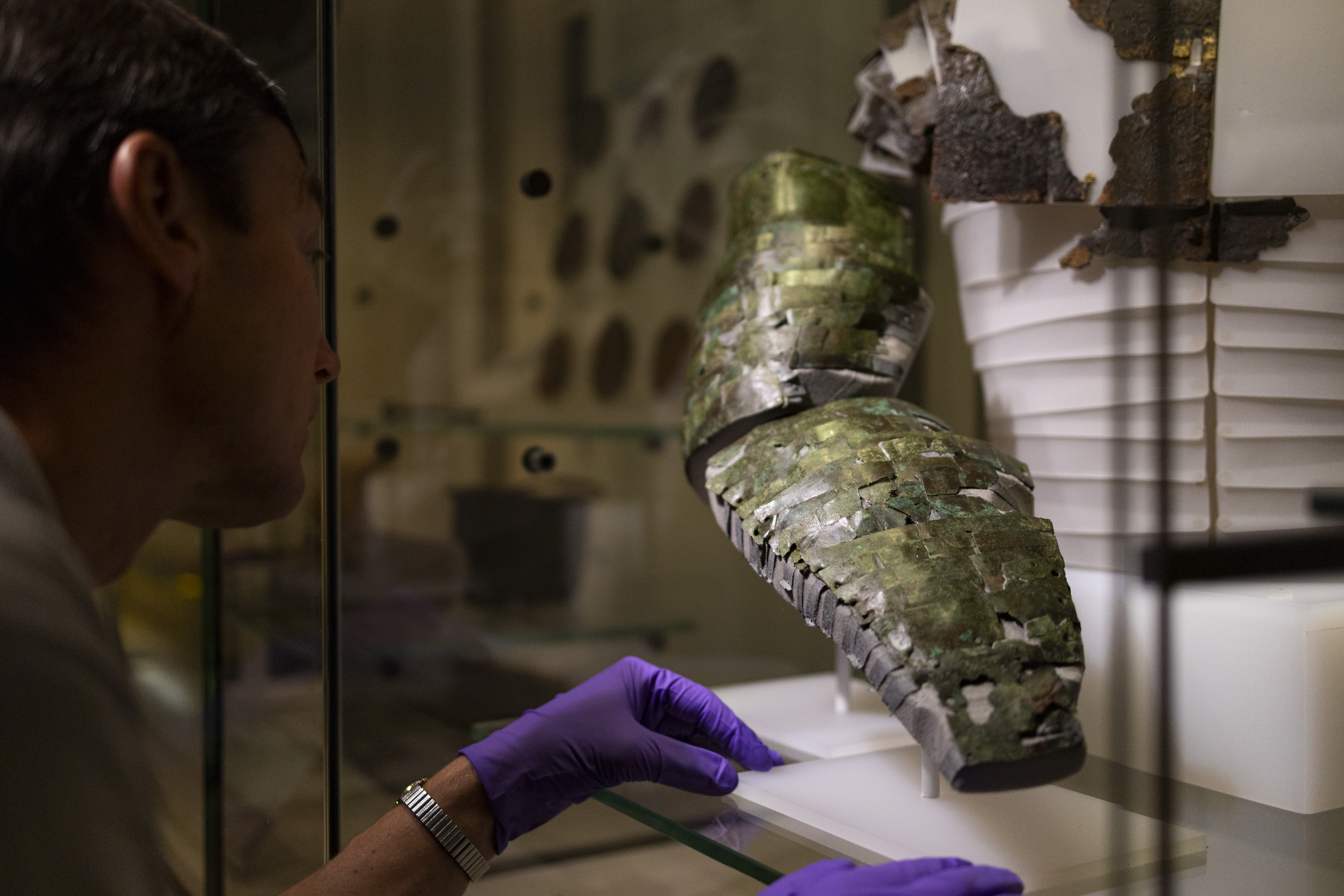 Close-up of the arm guard in a display case, with the purple-gloved hand of a member of museum staff visible next to it