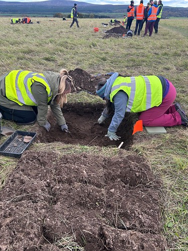 Two archaeologists dressed in high-vis jackets examining a square trench