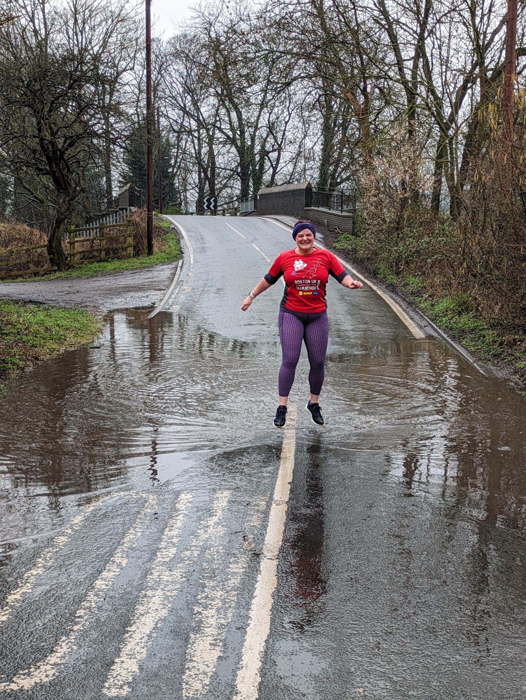 Stephanie Field smiling and jumping in a puddle while out running wearing a red top, purple leggings and and a blue hat 