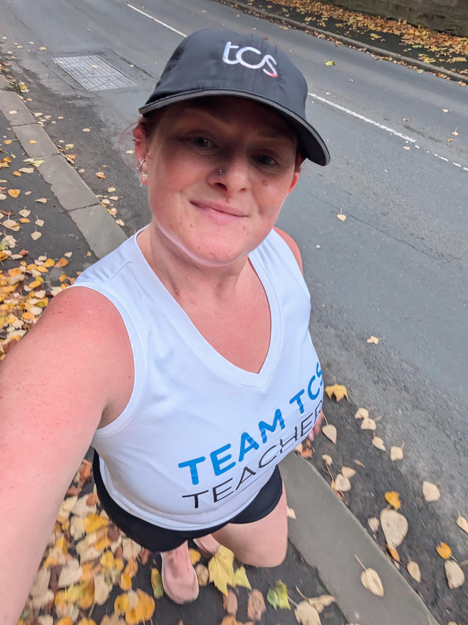 Stephanie Field looking at the camera while out running wearing a TCS cap, a white Team TCS Teachers vest and black shorts, surrounded by leaves on the pavement