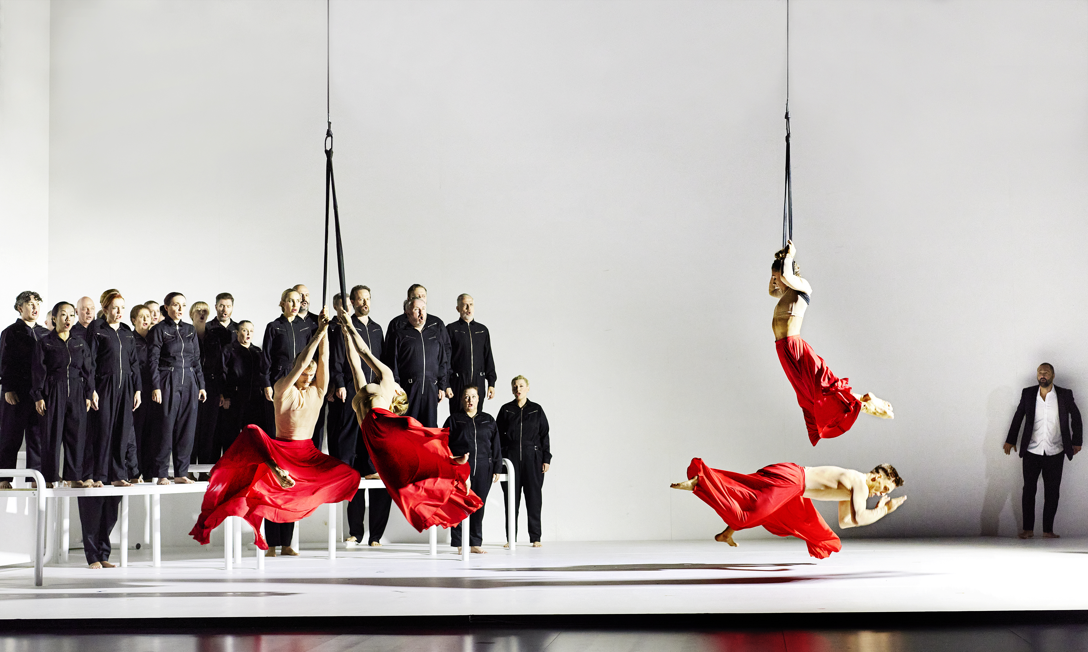 Acrobats dressed in red robes on a white stage with a choir dressed in black