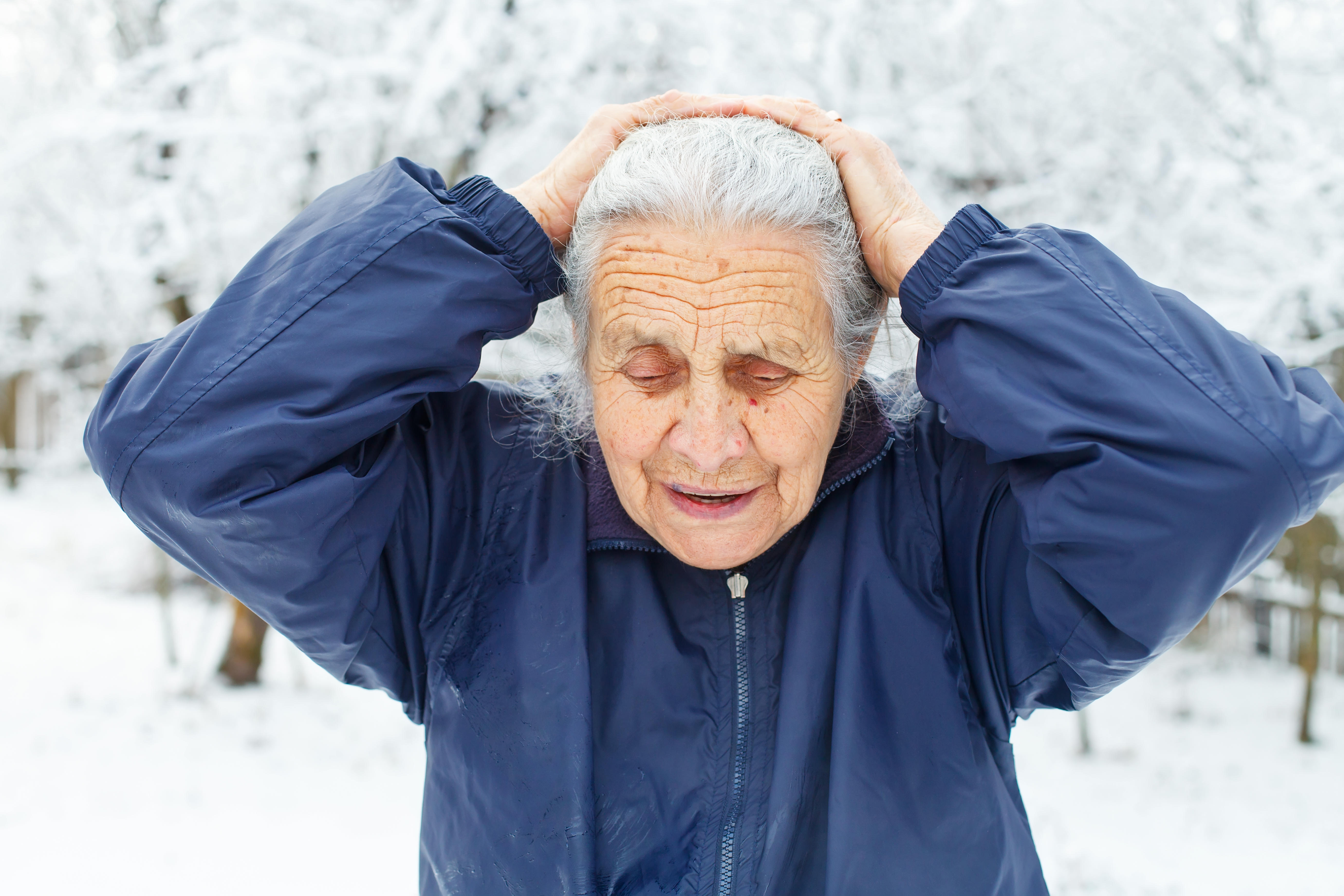 Picture of an old woman stood in a snowy park experiencing a headache 