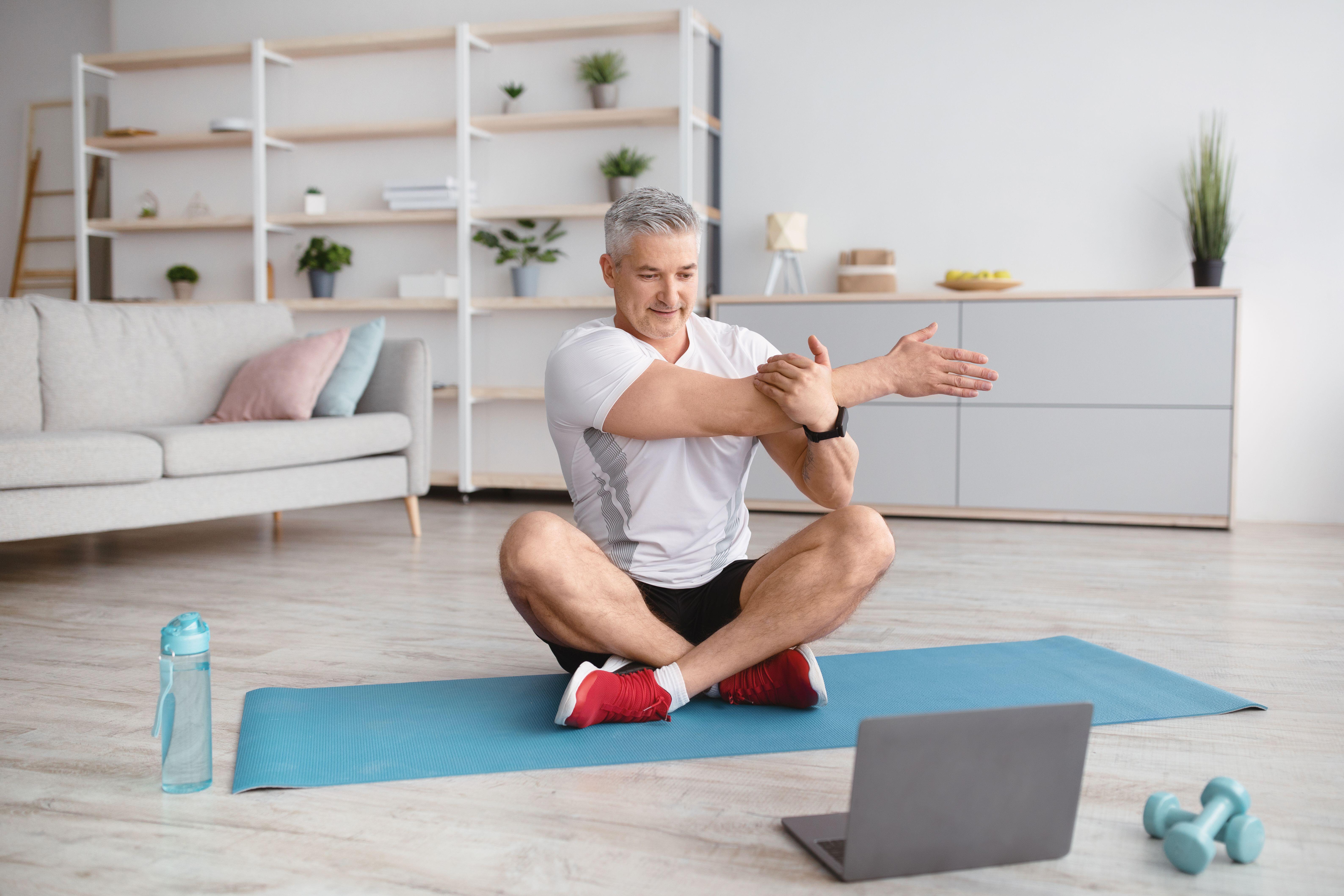 Athletic mature man doing yoga in front of laptop during home workout, practicing pilates, stretching on mat indoors