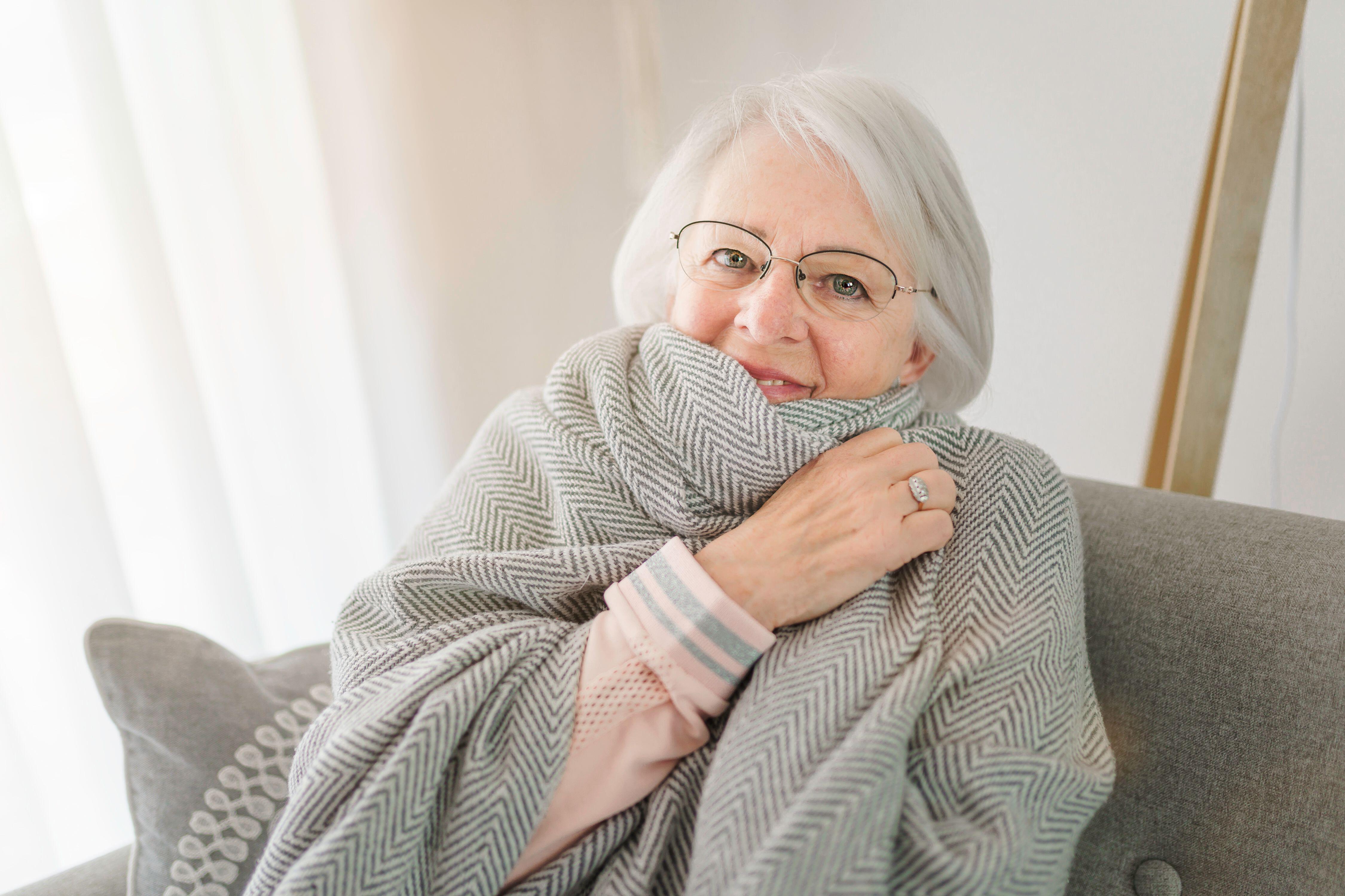 A picture of an elderly woman sitting on a sofa at home with wrapped in a grey blanket