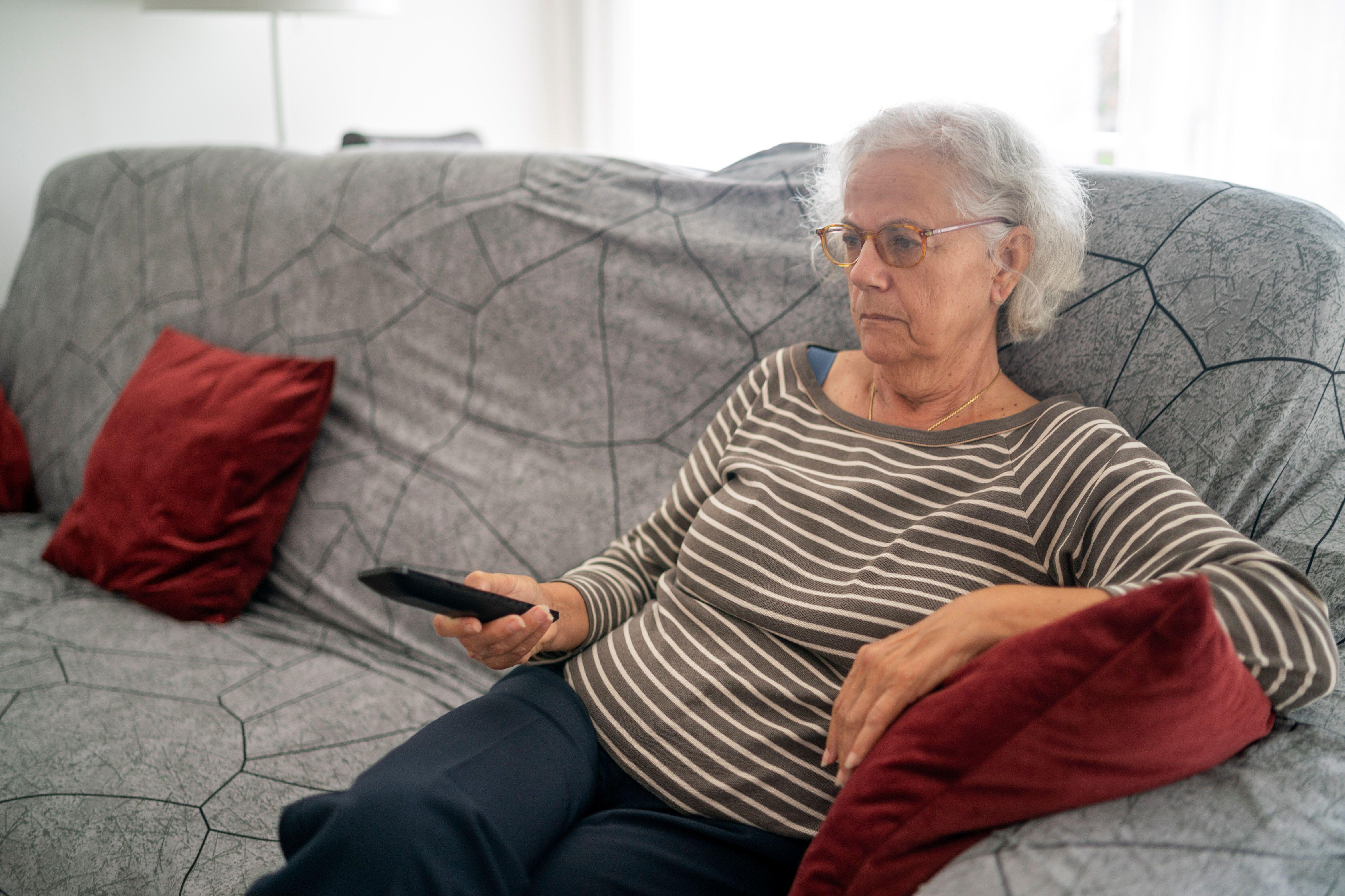 Senior woman sat on a grey sofa holding a TV remote