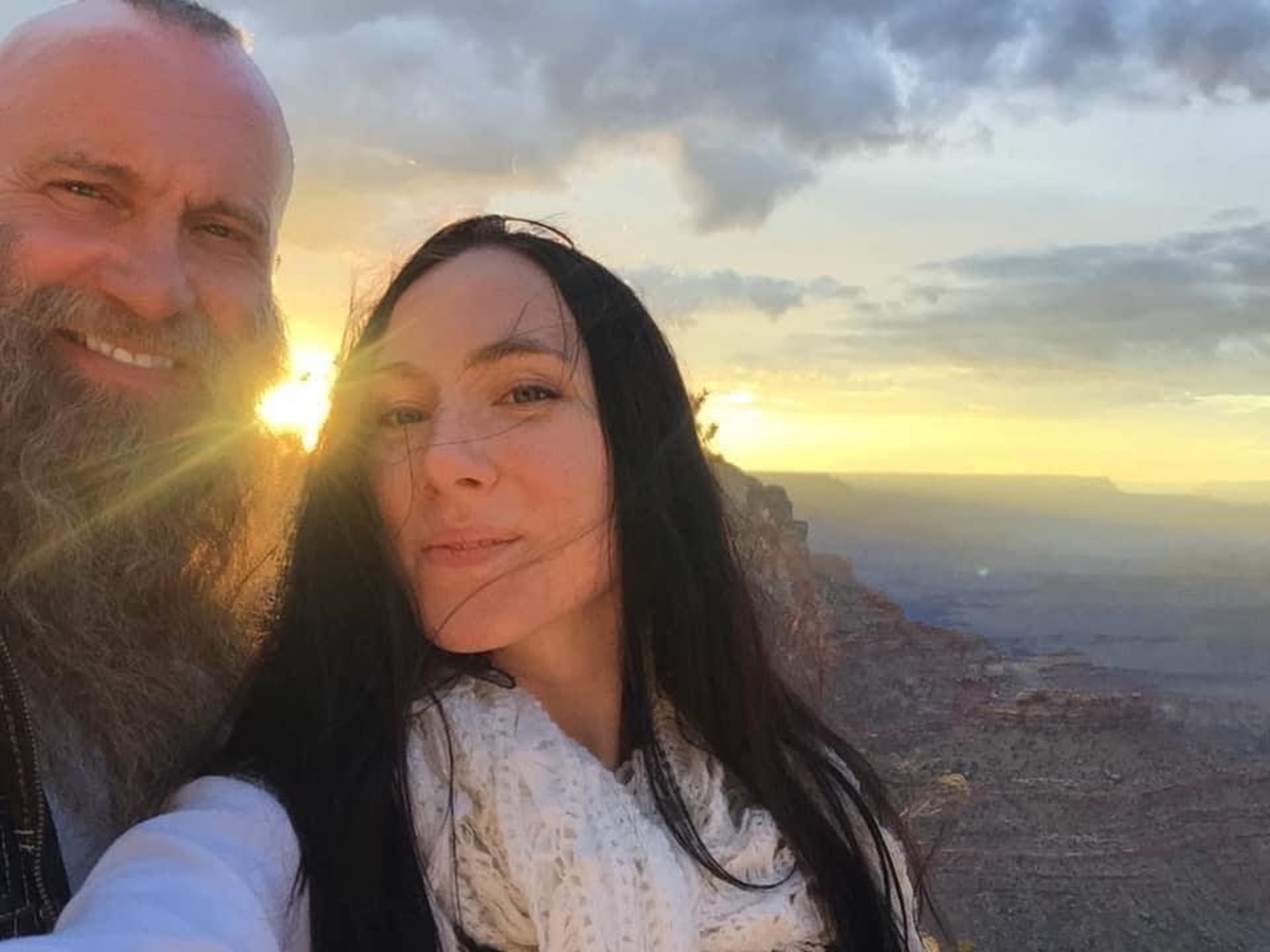 Charlie Allan and his wife, Chara Gallos-Allan, smile in front of a cloudy sky with sunlight shining through