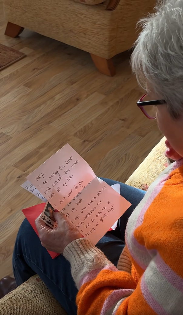 An elderly woman sat on her sofa and looking down at a handwritten letter