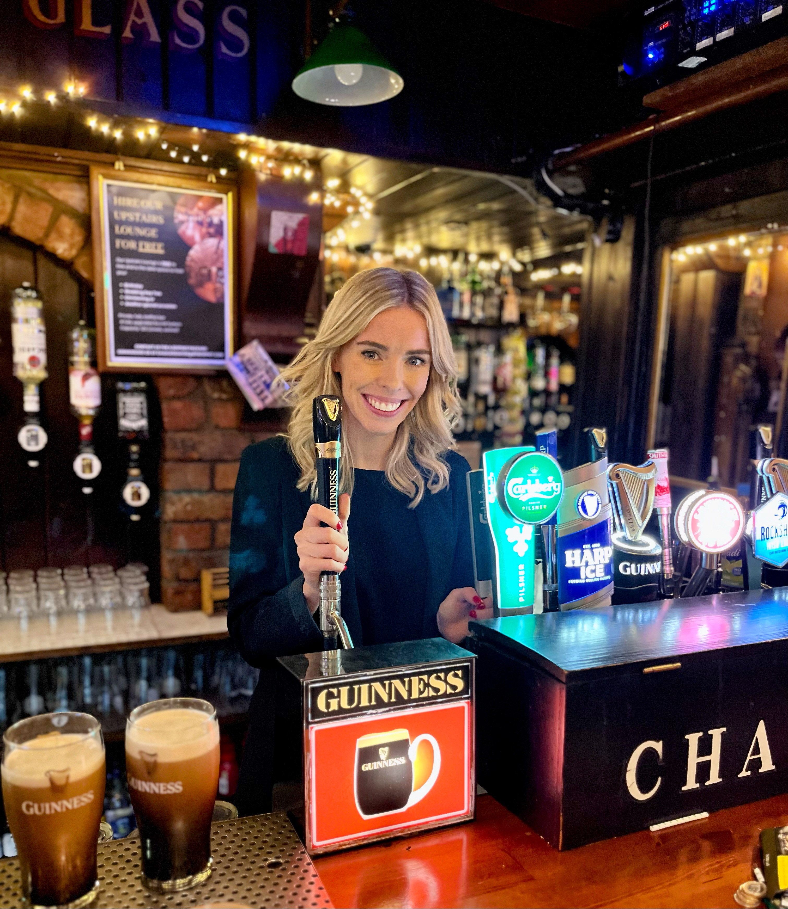 A blonde woman smiling while pulling a pint behind the bar of an Irish pub