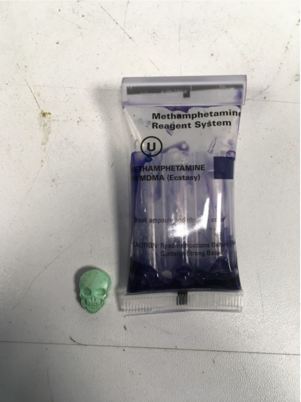 Green skull-shaped tablet next to purple coloured liquid in plastic testing bag