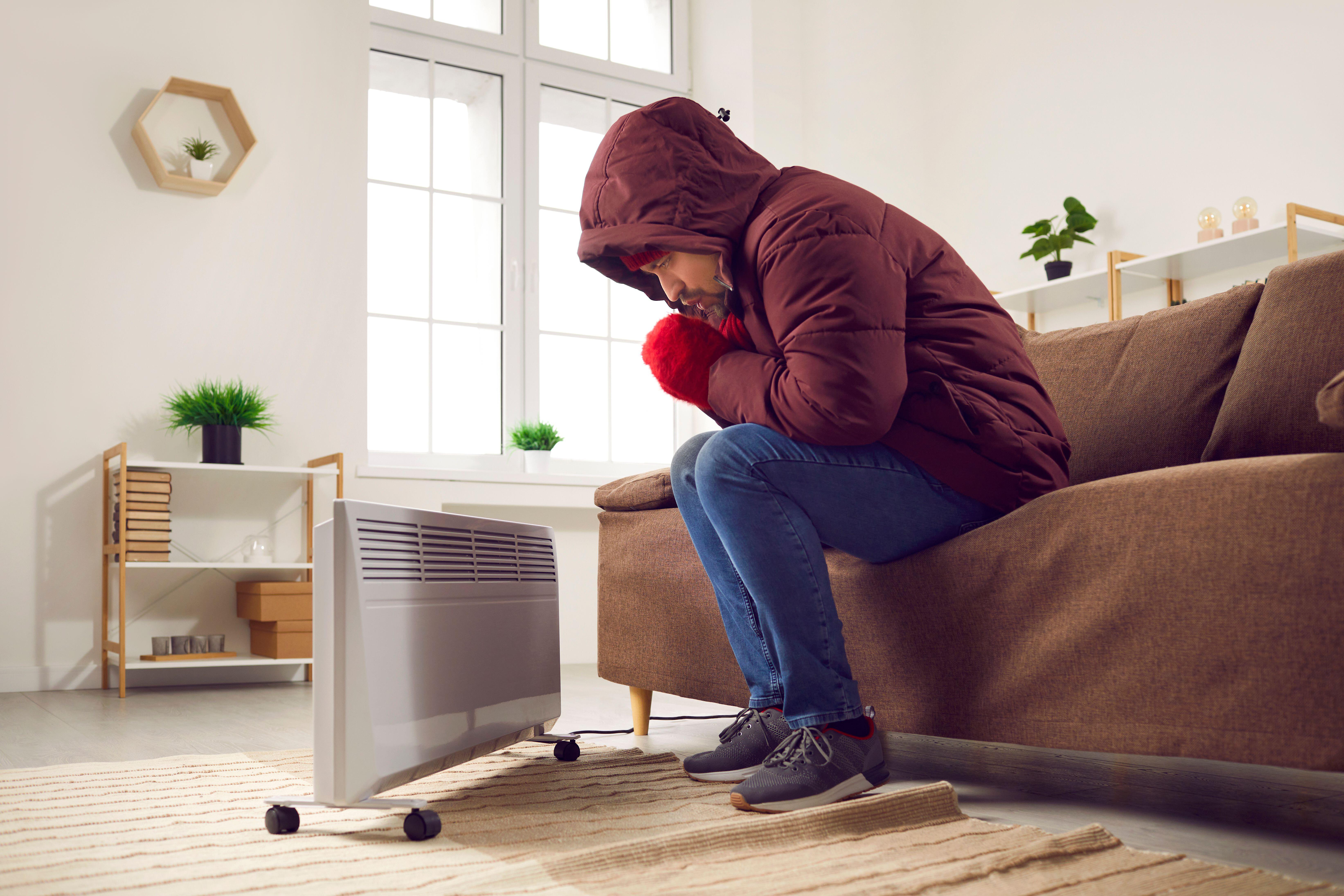 Man dressed in winter coat and gloves warming hands by an electric heater 