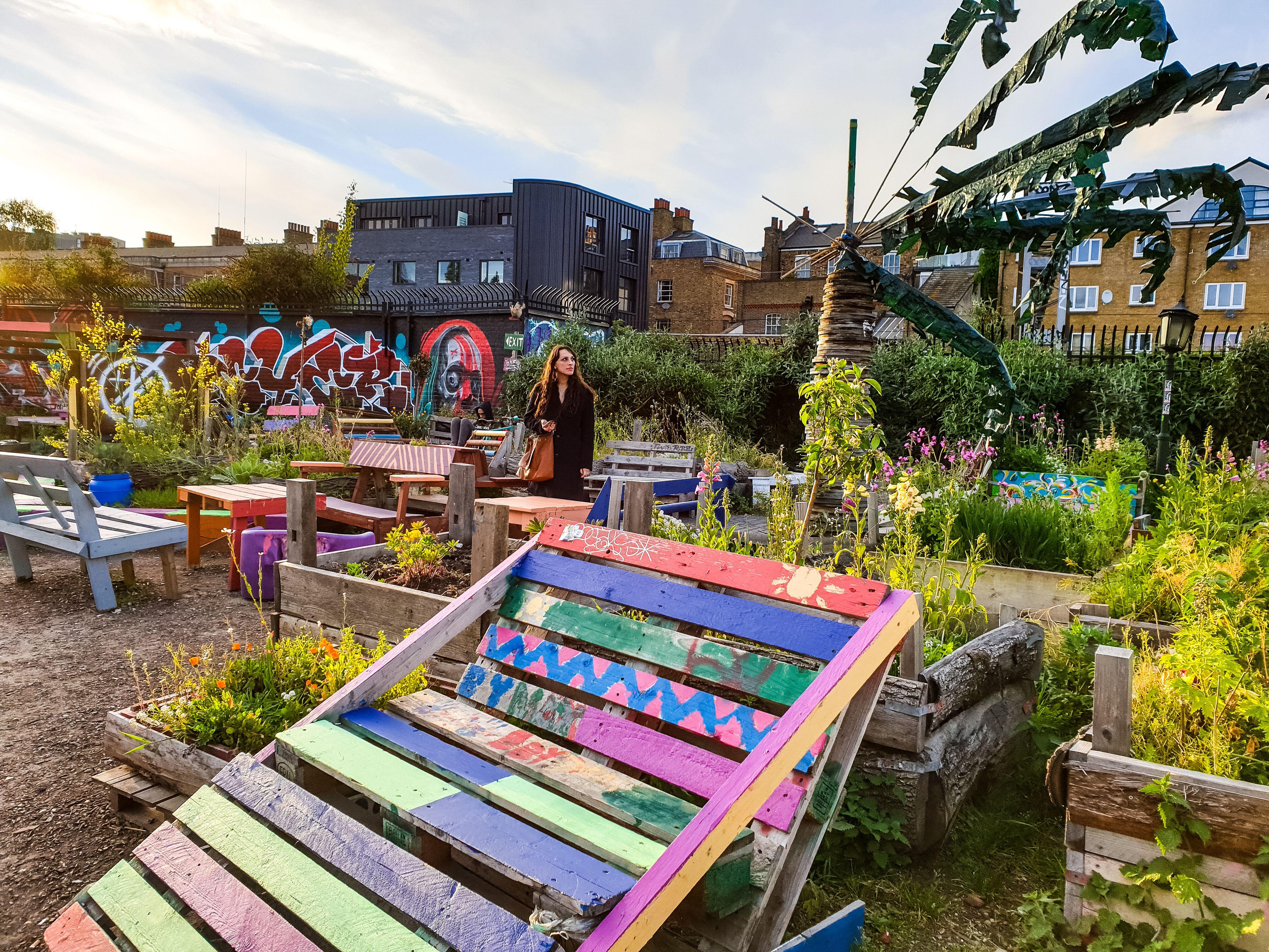 A woman standing in a community garden of raised beds and brightly painted furniture with buildings behind