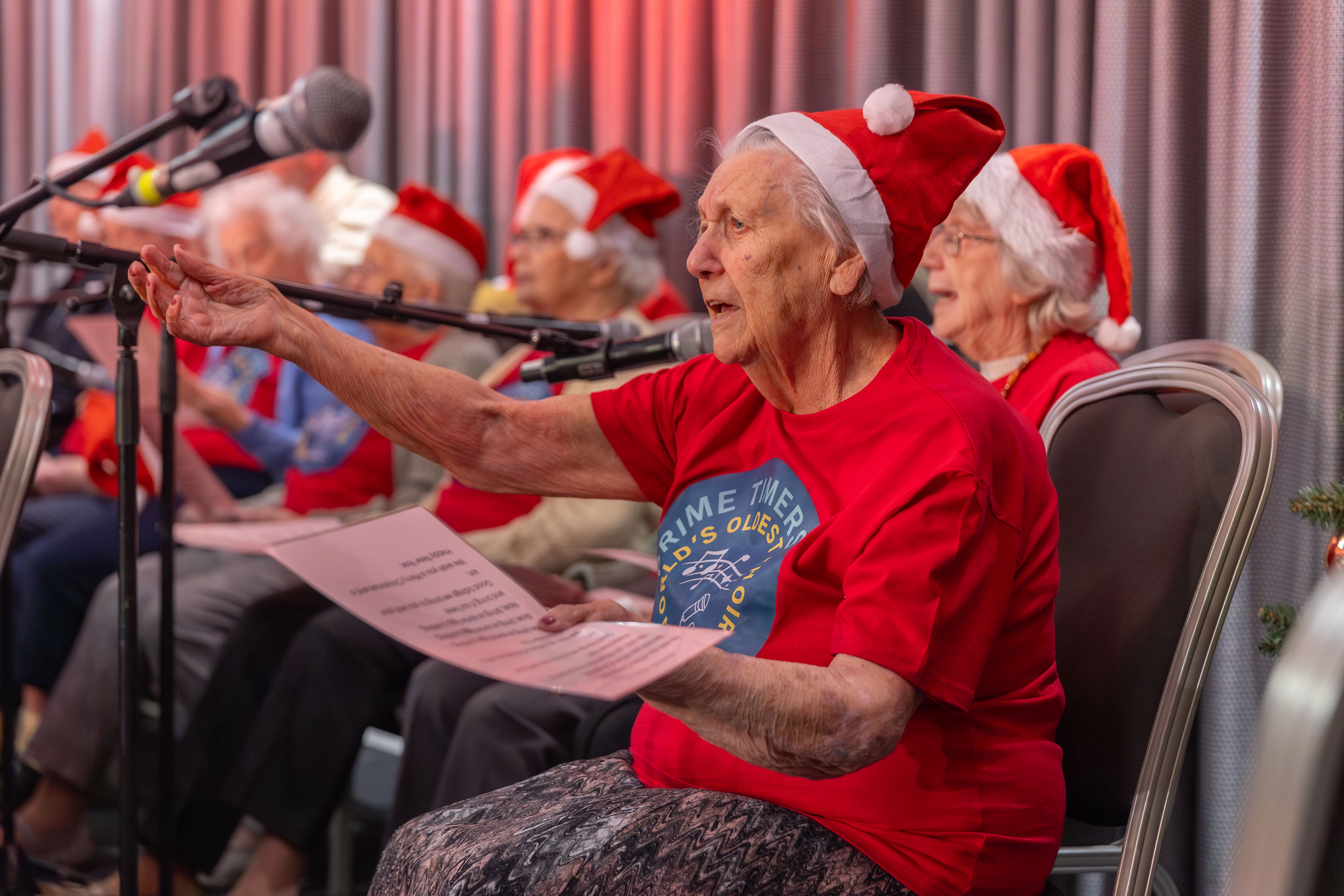 Elderly woman dressed in red top and Santa hat sat down and signing in front of a microphone