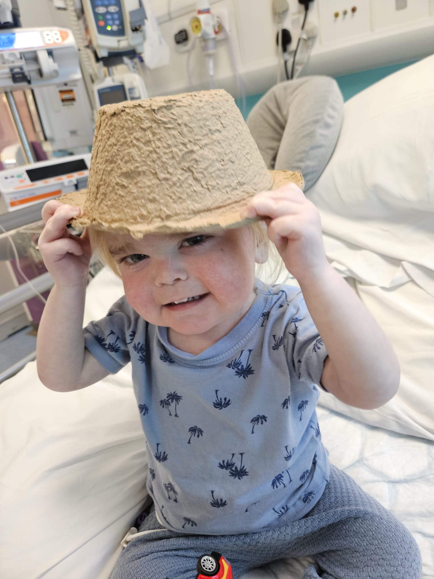 Alfie sat on a hospital bed pretending a cardboard waste bowl is a hat